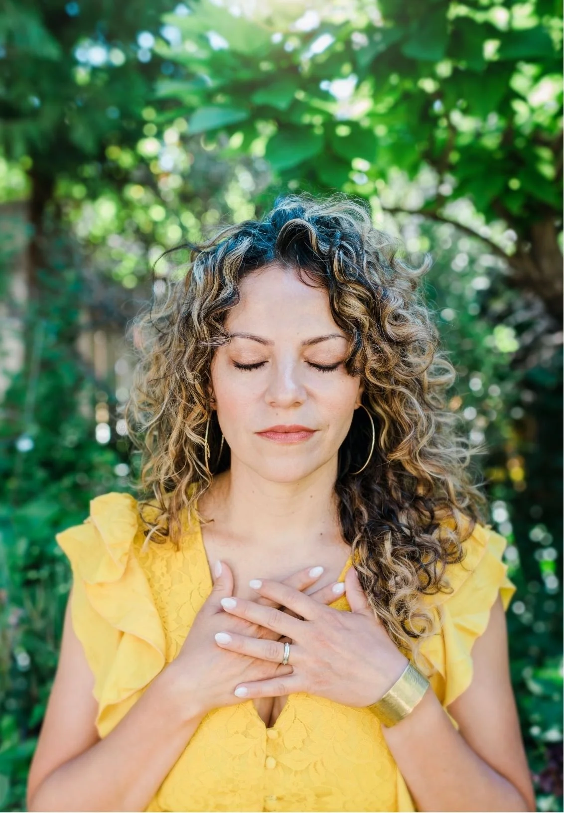 Mery Vargas Mares, therapist in Reno, NV with curly hair wearing a yellow dress, with her eyes closed and hands on her chest in a garden.