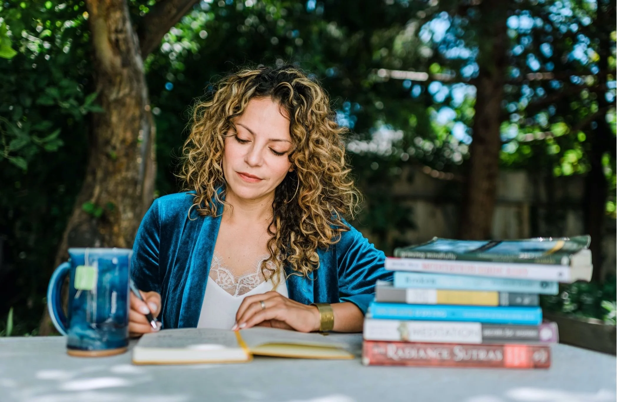Mery Vargas Mares, therapist in Reno, NV, with curly hair wearing a blue jacket sitting at a table outdoors, reading a book with a stack of books nearby.