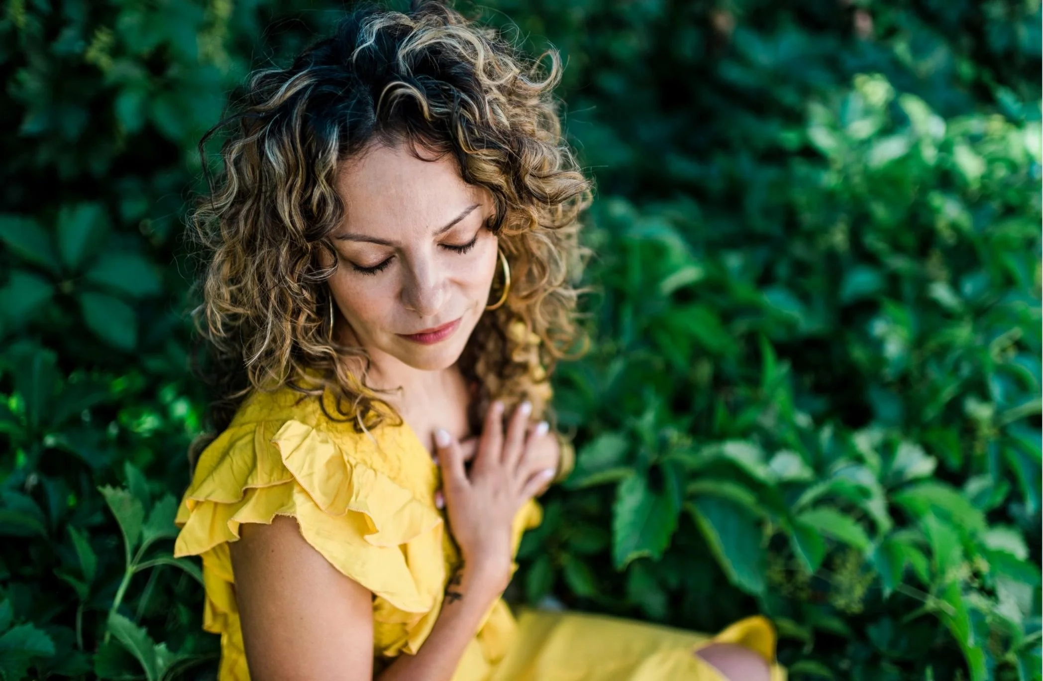 Mery Vargas Mares, therapist in Reno, NV, with curly hair and gold hoop earrings in a yellow dress, eyes closed, with her hand on her chest, standing in front of lush green foliage.