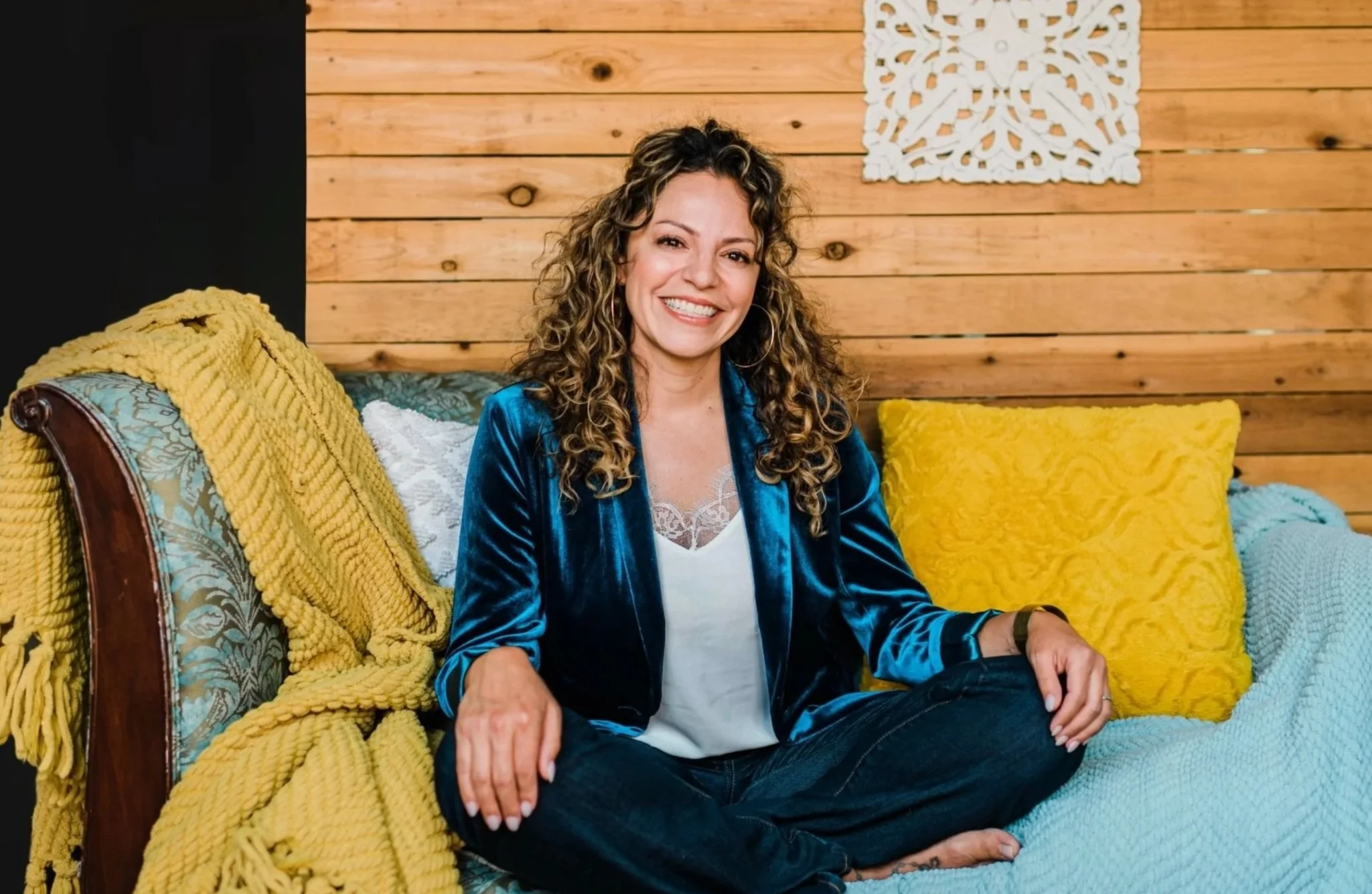 Mery Vargas Mares, therapist in Reno, NV smiling with curly hair, sitting on a patterned sofa with a yellow blanket and yellow pillow in a room with wooden walls and a decorative white wall art piece.