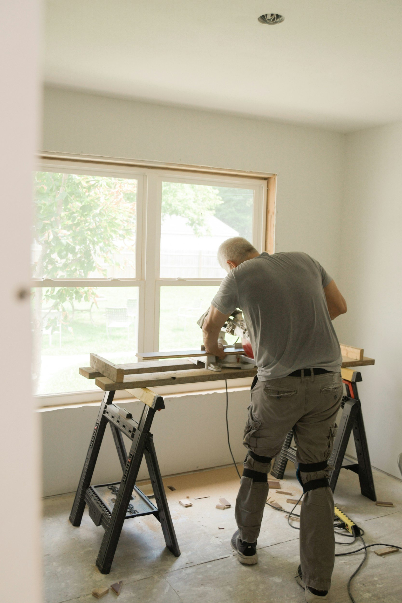 A person is working on a home improvement project inside a room with a large window. They are standing on a step ladder and using a saw on a wooden board secured on a workbench. The room is brightly lit by natural light, and there are some tools and wood pieces on the floor.