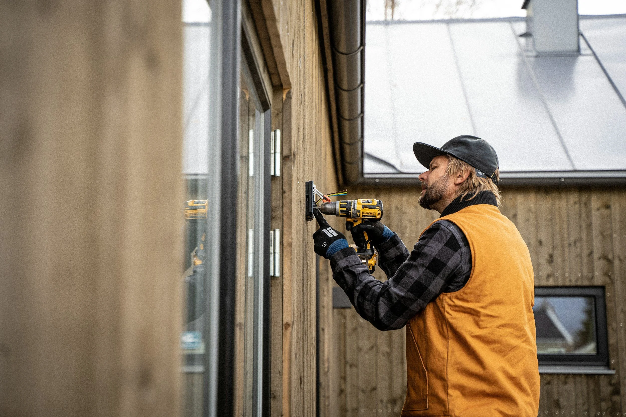 A man wearing a black cap and orange vest using a power drill on a wall in a building under construction or renovation.