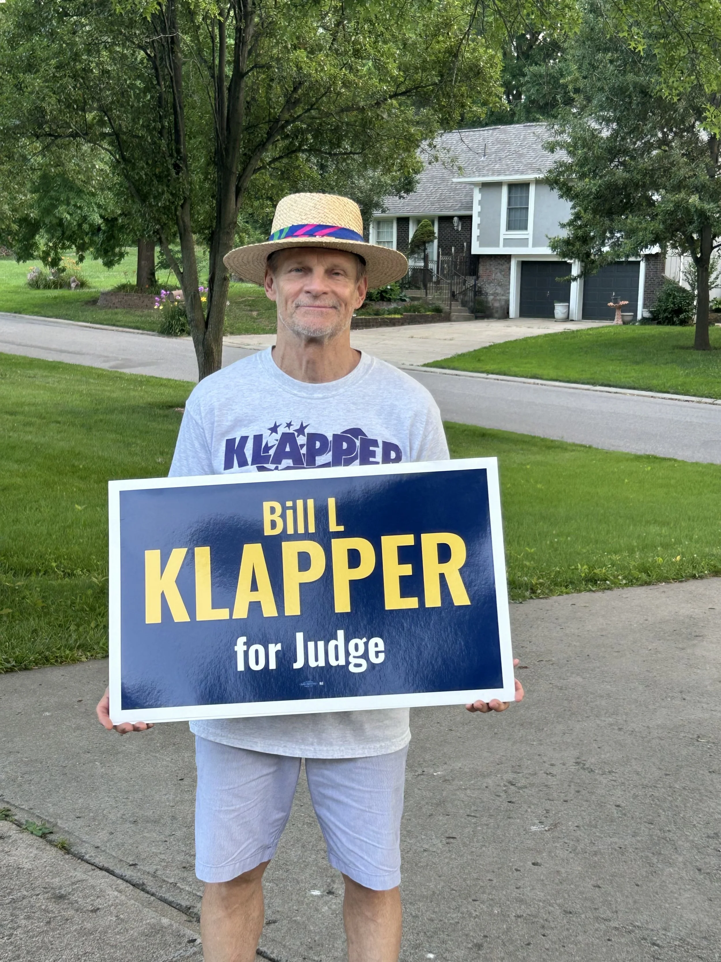 Judge Klapper holding a yard sign that reads 'Bill L Klapper for judge.'  He is wearing a light gray T-shirt and shorts, and a wide-brimmed straw hat with a colorful band. Behind him is a residential street.