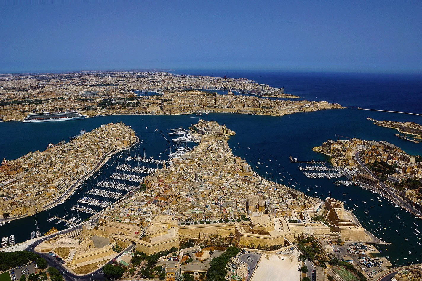 An aerial view of Valletta, Malta, showcasing the old fortified city with historic buildings, a harbor filled with boats, the Mediterranean Sea, and surrounding modern cityscape on a sunny day.