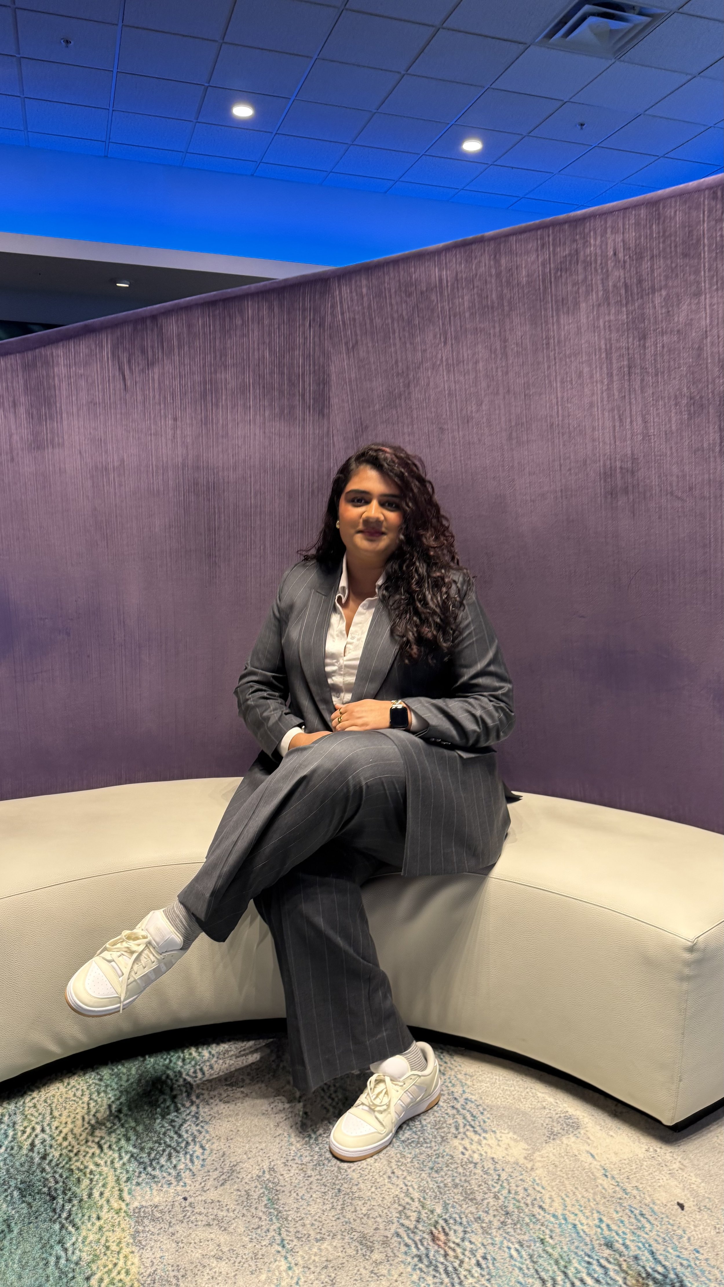 A woman with curly dark hair in a gray pinstripe suit sitting on a beige curved sofa in a modern room with purple and blue lighting.