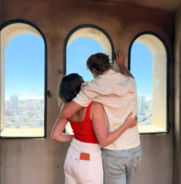 A man and woman stand, embracing, in front of three arched windows overlooking a city on a sunny day. 