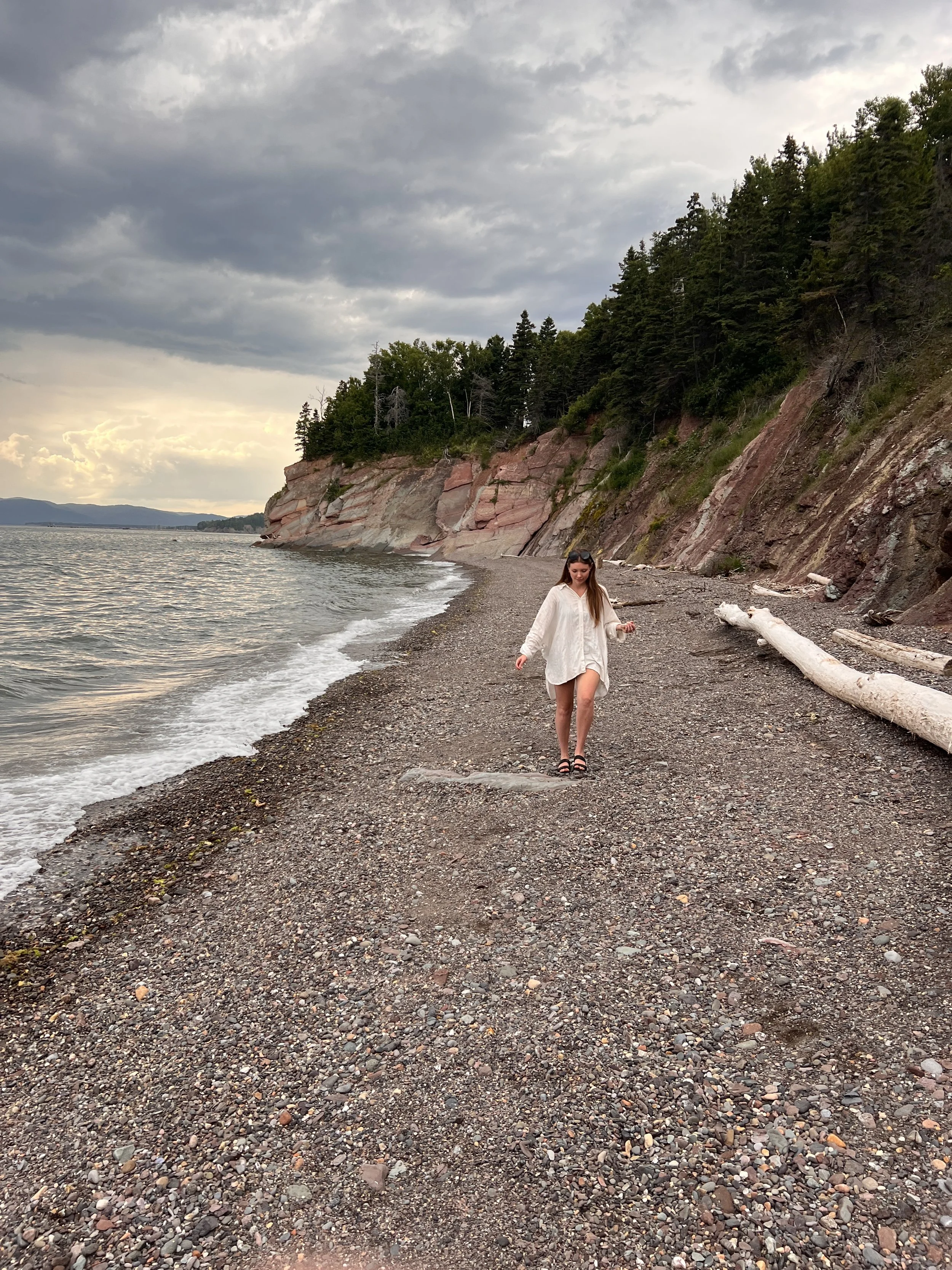 A woman wearing a white shirt walking barefoot on a rocky beach near a forested cliff with mostly cloudy skies.