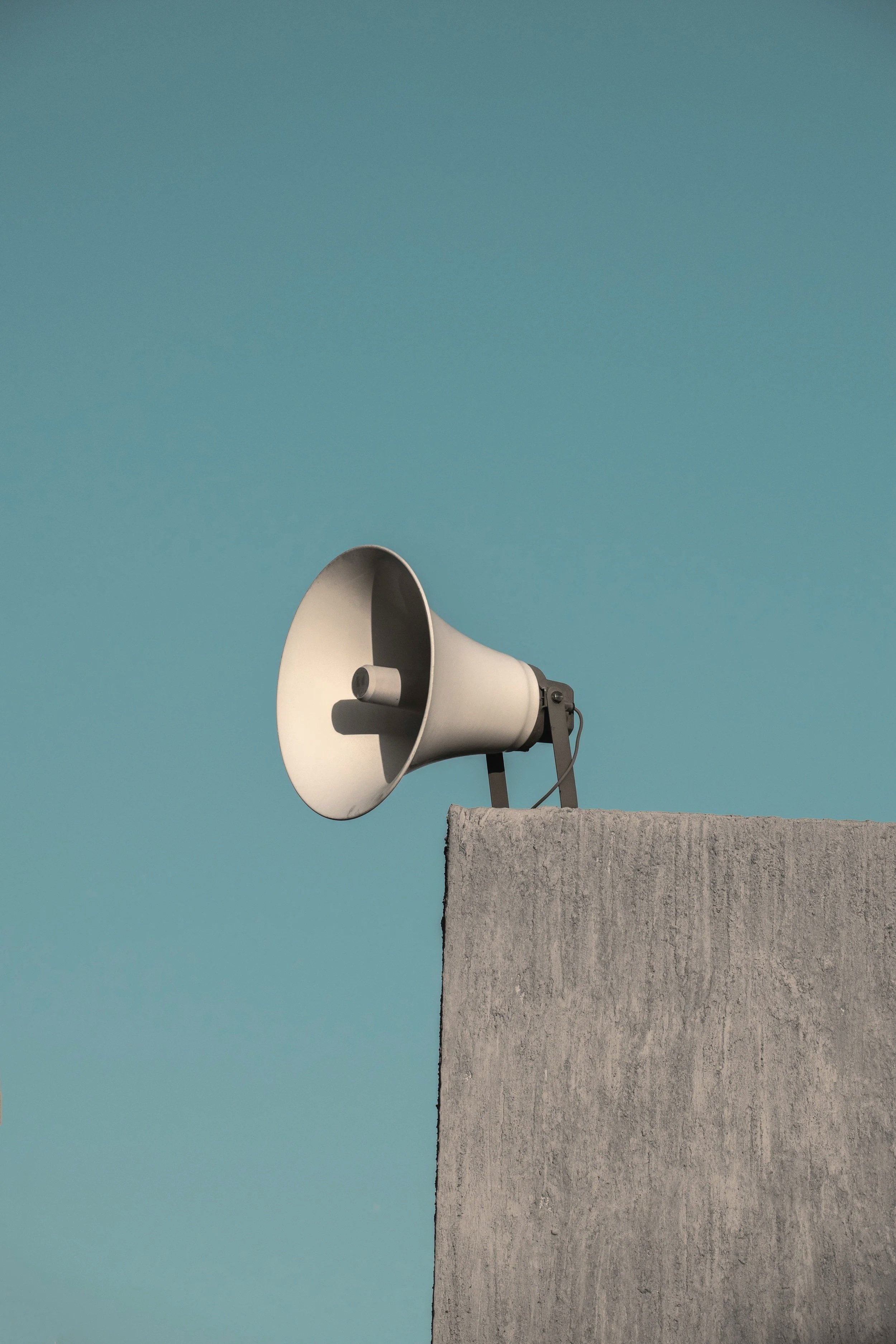 A white loudspeaker mounted on a gray concrete wall against a clear blue sky.