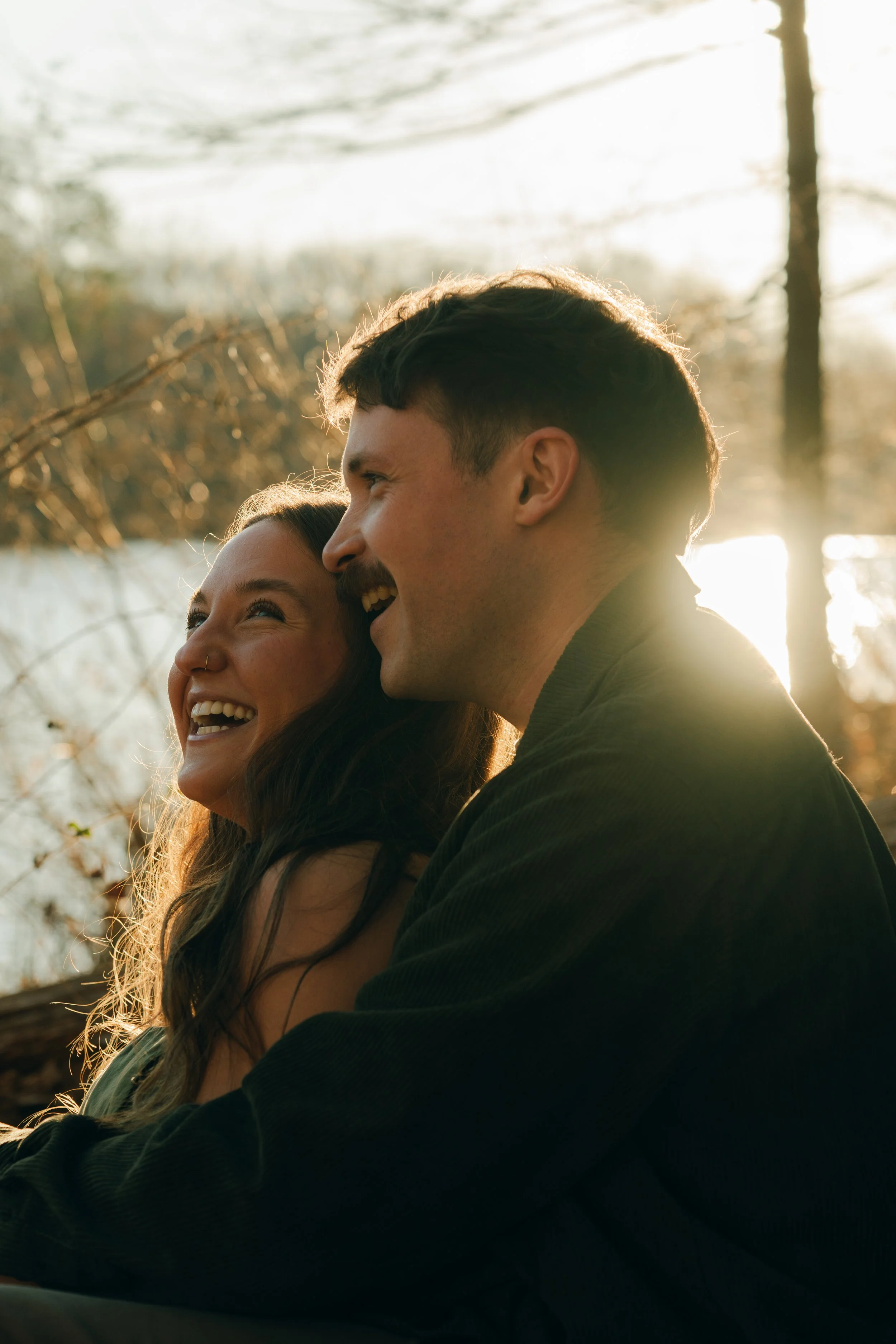 A joyful couple, a woman and a man, smiling and cuddling outdoors during sunset with trees and a body of water in the background.