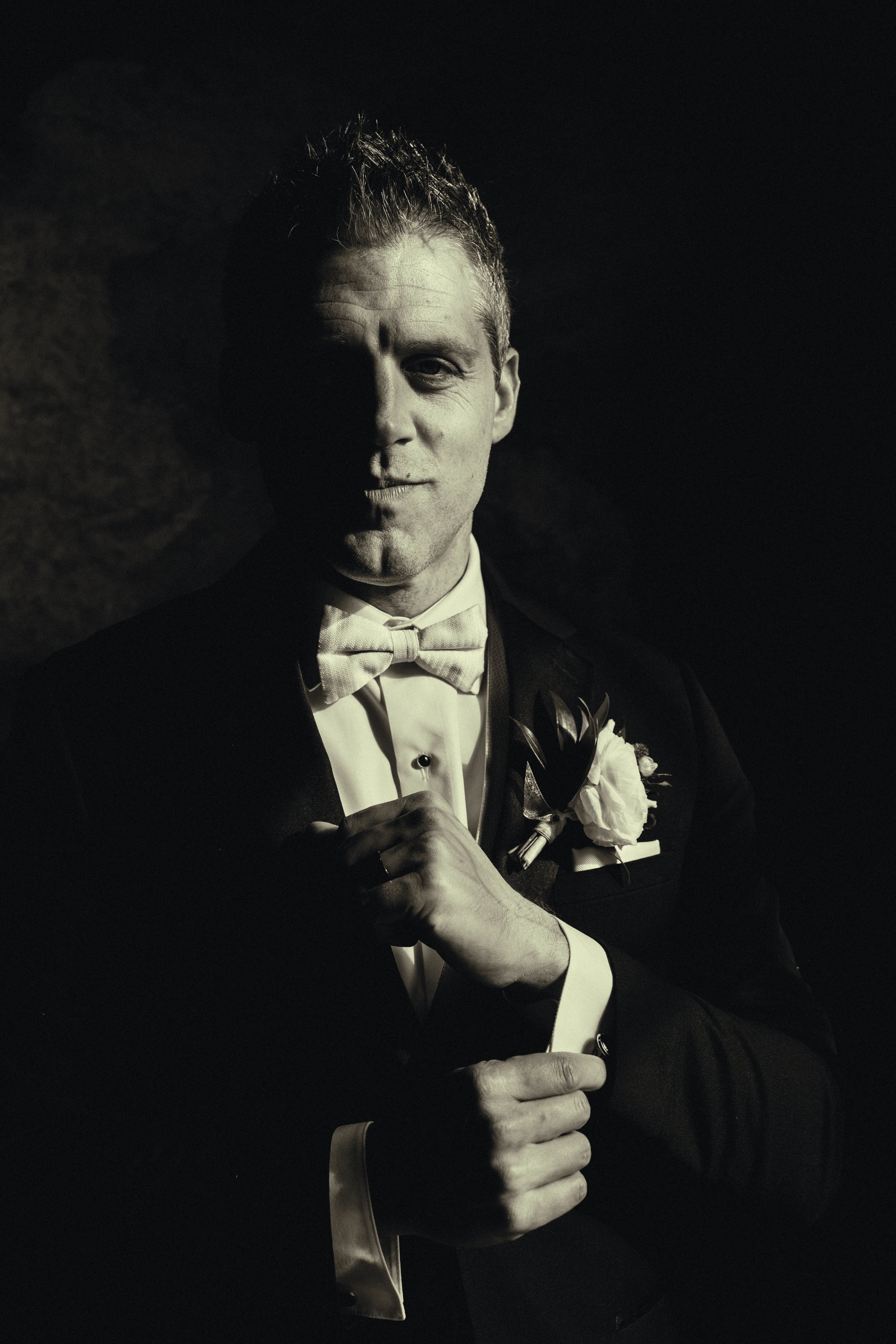 A black and white portrait of a man dressed in formal attire, including a tuxedo, bow tie, and boutonniere, smiling subtly and standing against a dark background.