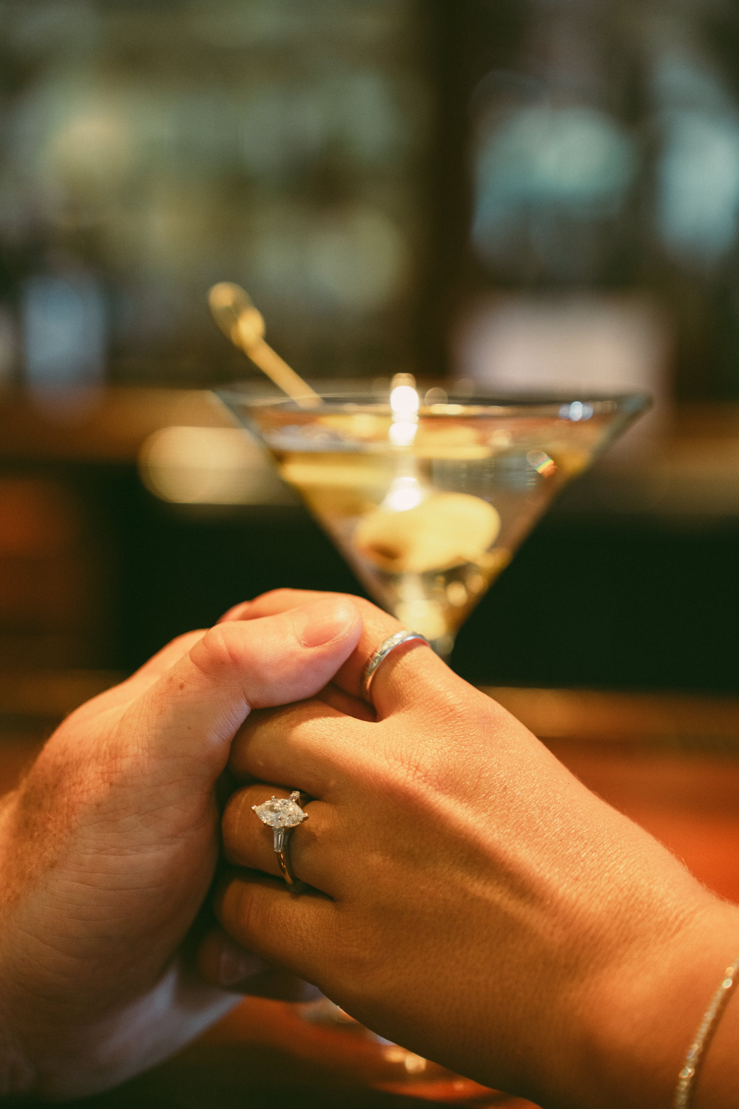 A person holding a martini glass with a clear liquid and an olive garnish. The person is wearing a diamond ring on their left hand, with a smaller ring on the pinky finger. The background is blurred with soft lighting.