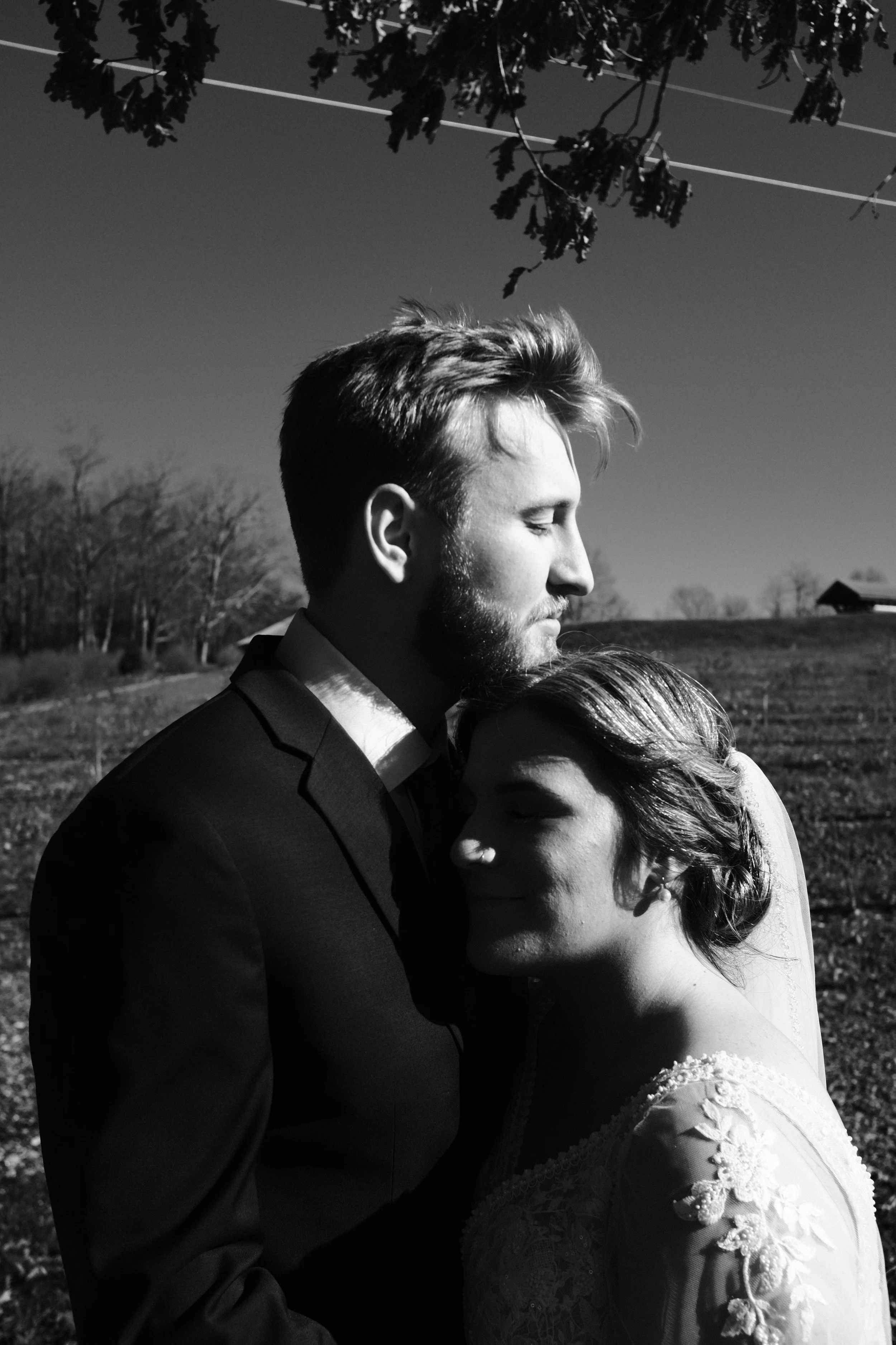 Black and white photo of a bride and groom embracing outdoors. The groom has a beard, short hair, and is wearing a suit. The bride has shoulder-length hair with curls, wearing a lace dress with floral embroidery, and her eyes are closed as she presse
