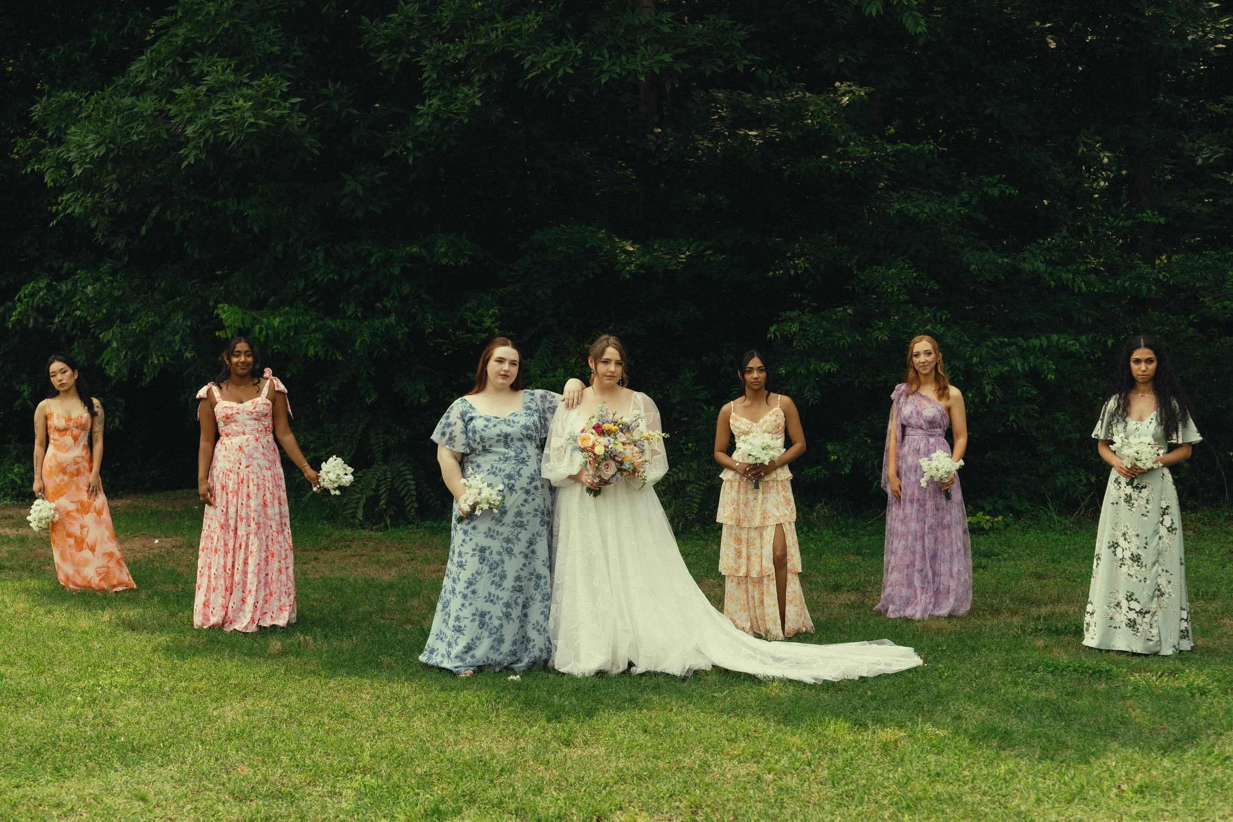 A bride and six women in colorful dresses standing outdoors on grass, holding bouquets of flowers, with trees in the background.