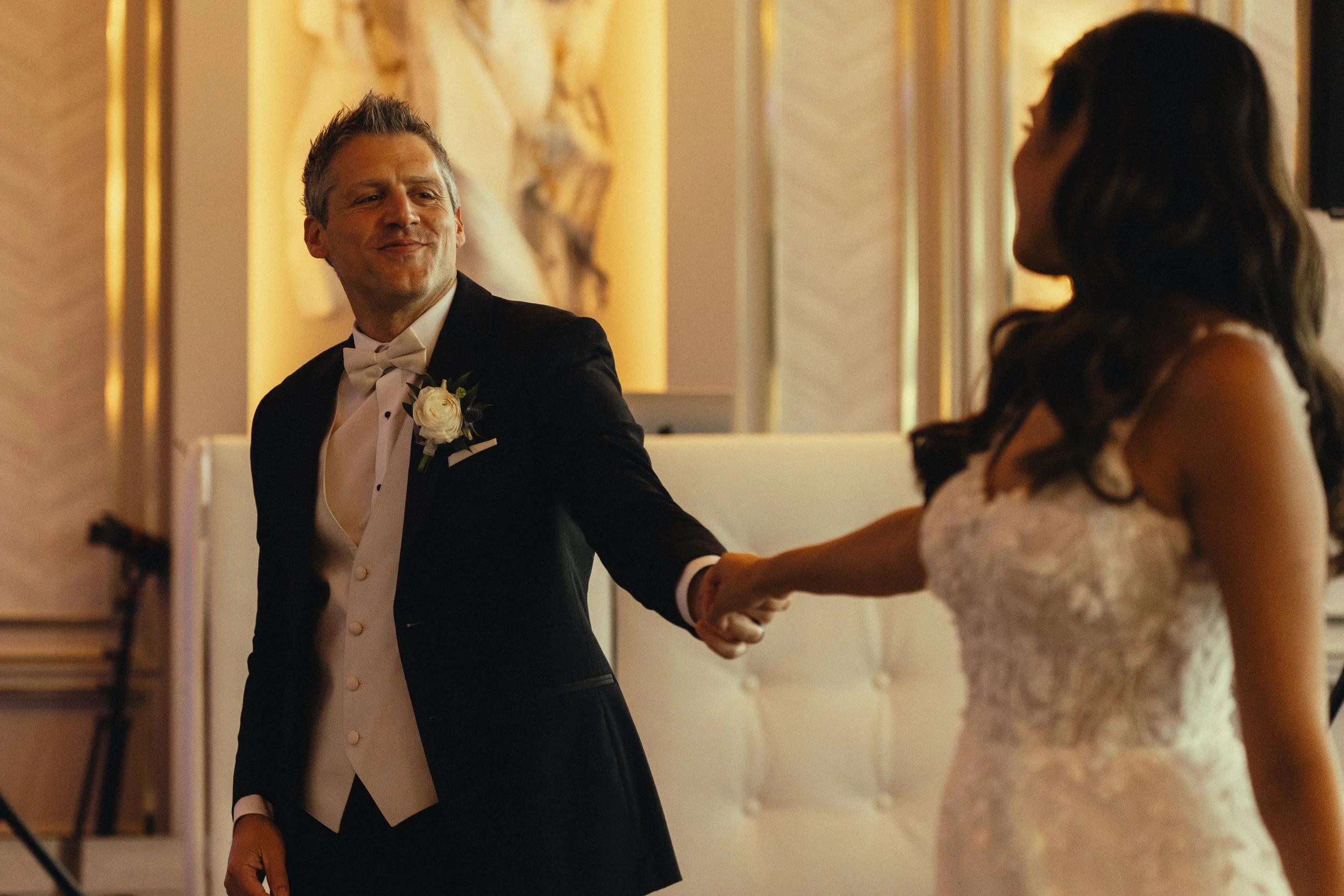 A man in a tuxedo with a white bow tie and boutonniere holding hands with a woman in a white lace wedding dress. They are dancing in a warmly lit room with ornate decor.