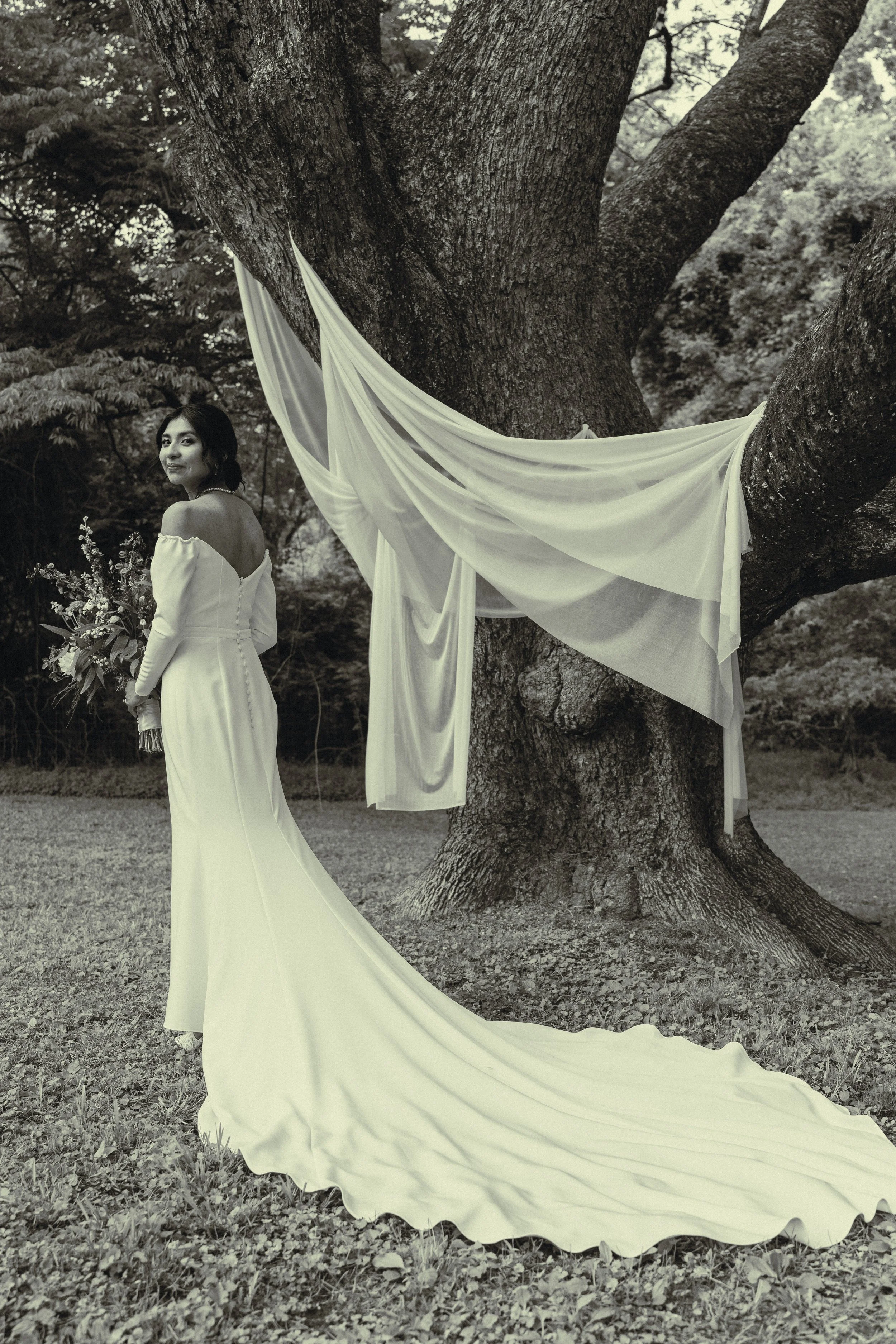 A woman in a wedding dress standing outdoors next to a large tree with a draped fabric decoration, holding a bouquet of flowers, in a black and white photograph.