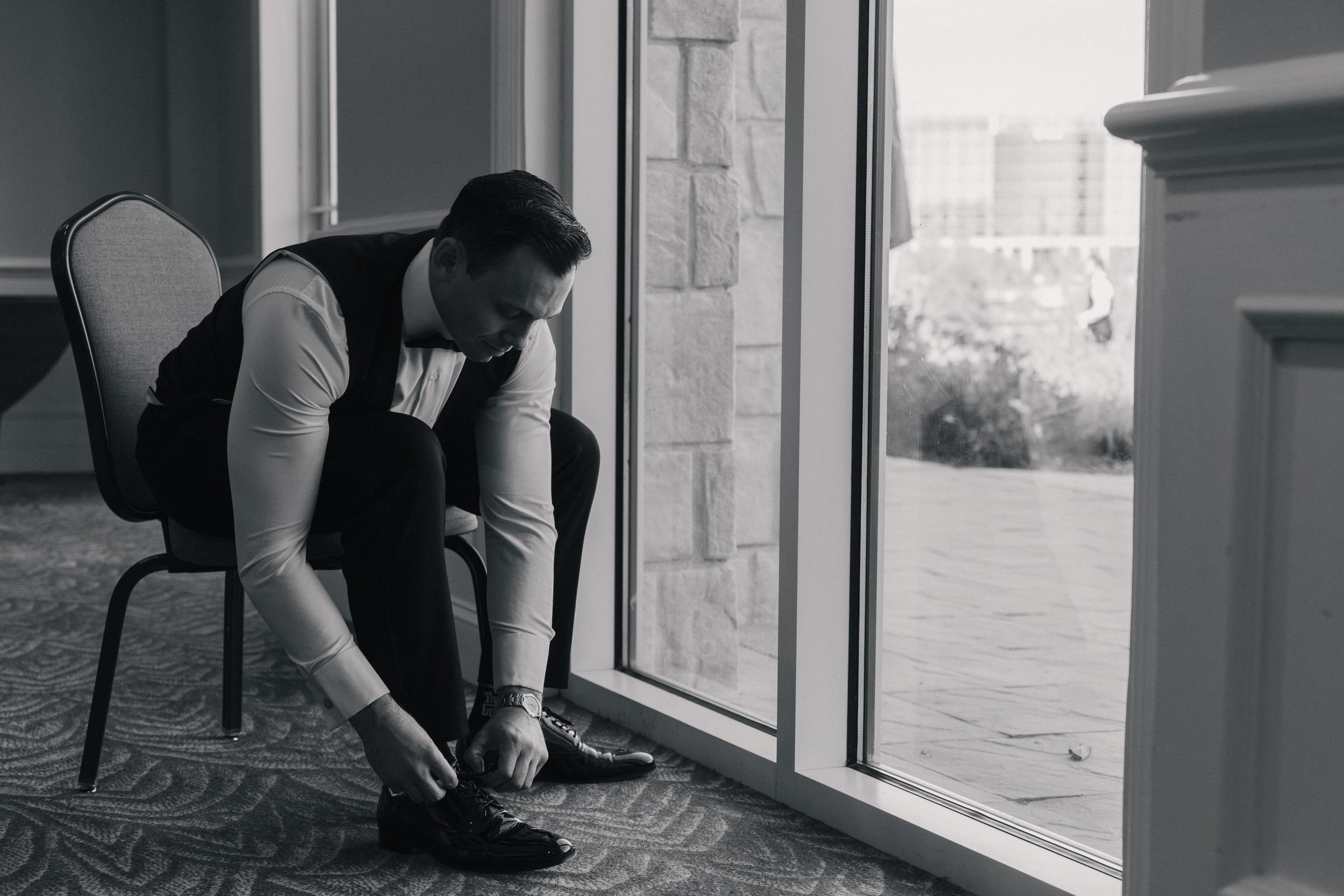 A man in formal attire, sitting in a waiting area, tying his shoelaces, near large windows overlooking an outdoor scene.