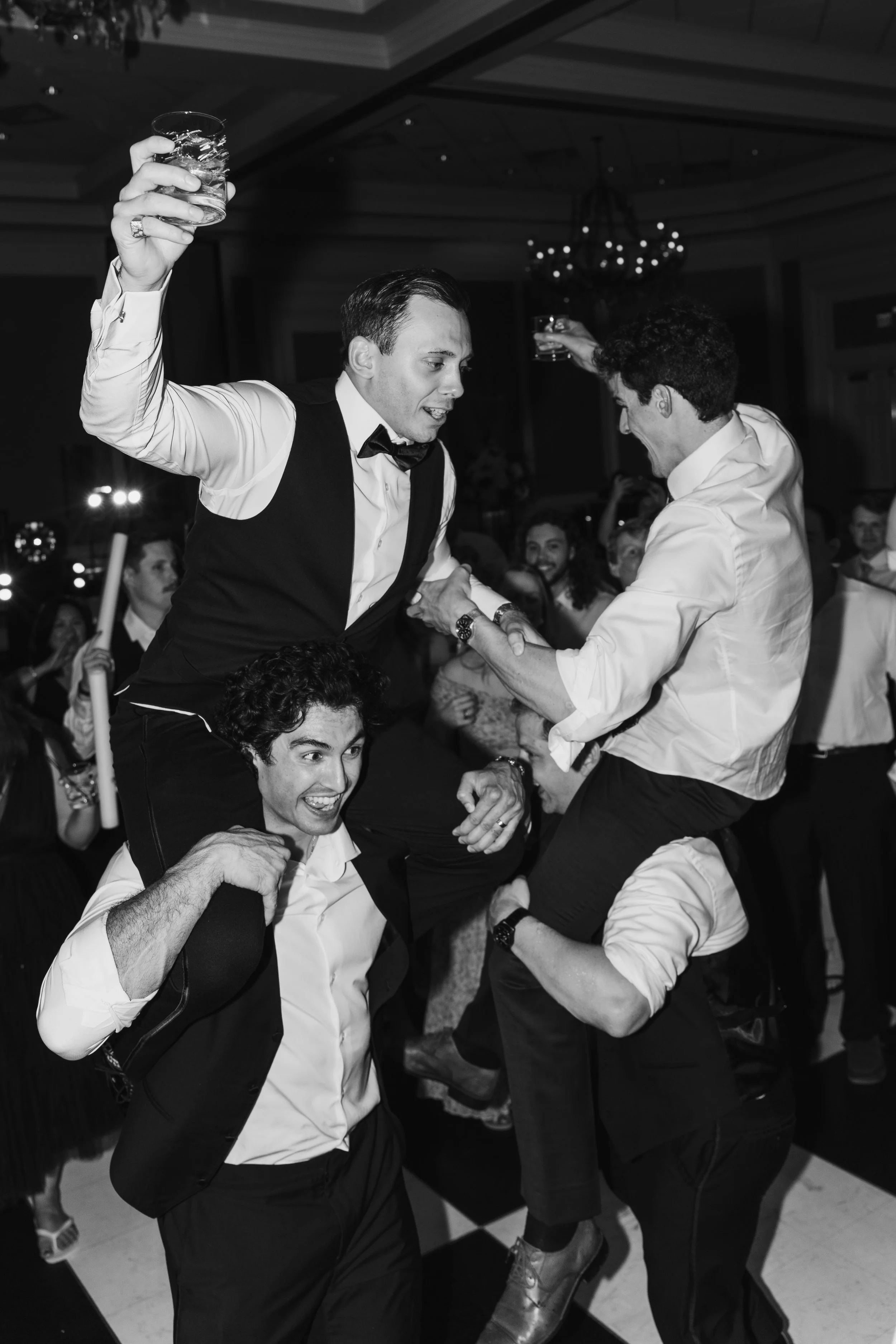 Two men are dancing and holding glasses at a celebration or party, with other guests smiling in the background.