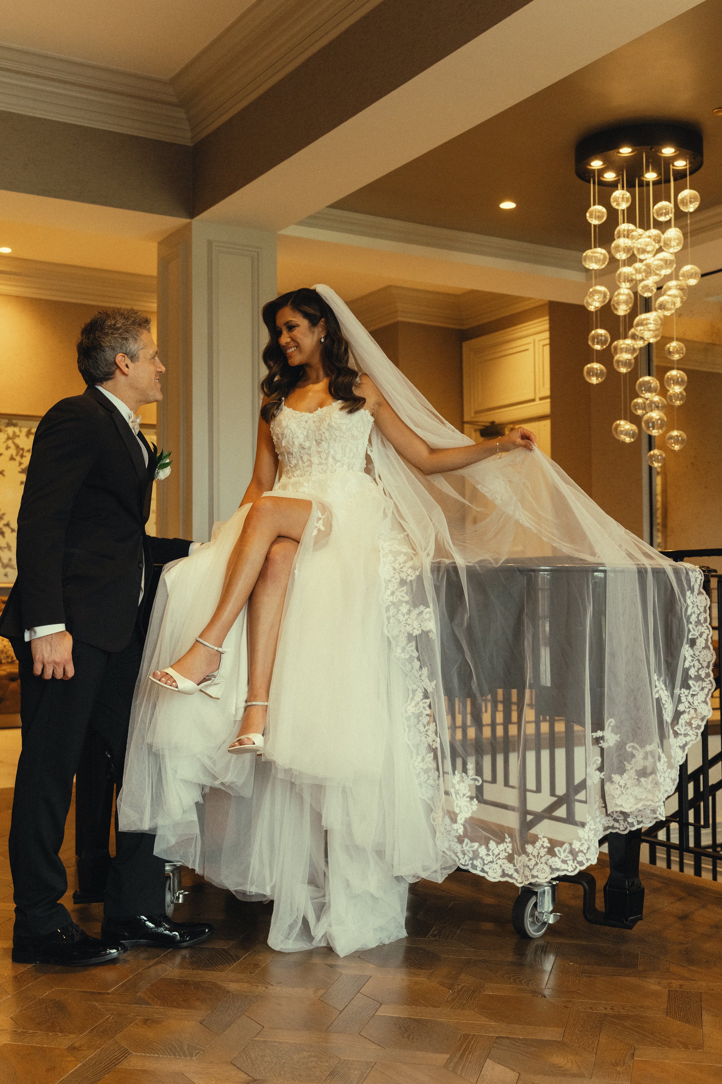 A bride with long dark wavy hair in a white wedding gown sitting on a table poseing with a big smile, holding her dress with one foot. A man in a black tuxedo with a boutonniere is smiling at her, holding her leg. The setting is a formal indoor venue