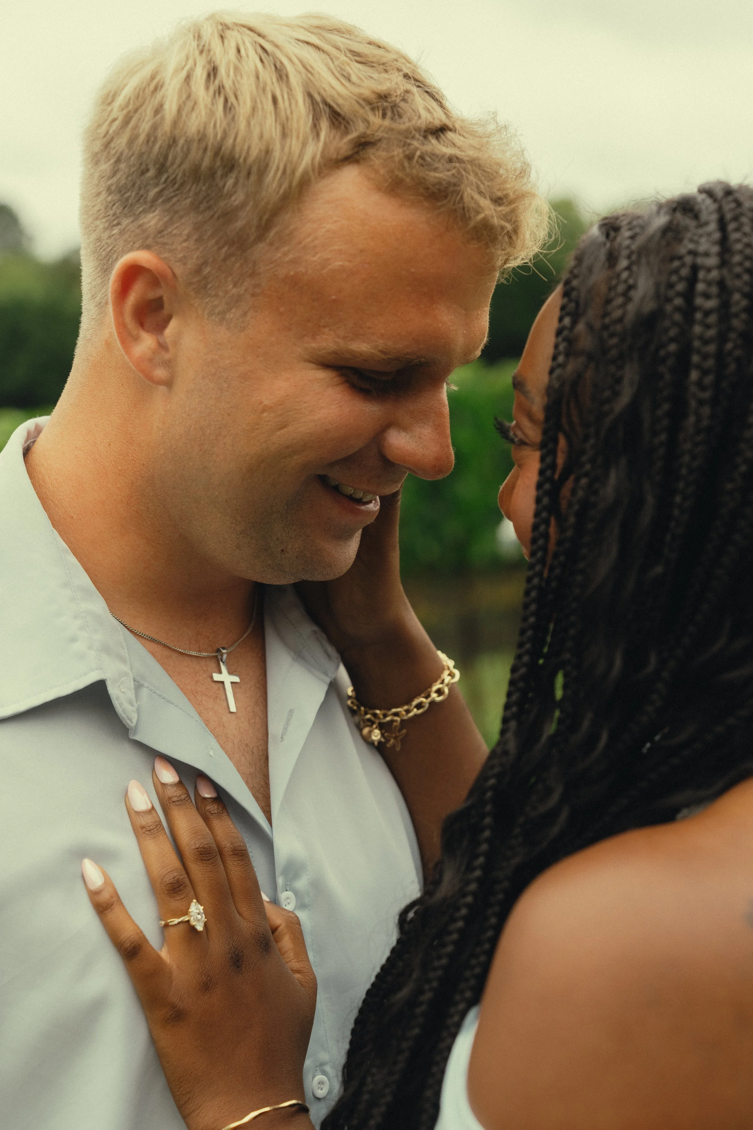 A couple is close to each other, smiling, with their foreheads touching. The woman has her hand on the man's chest, displaying jewelry including rings and a bracelet. The man is wearing a light-colored shirt with a cross necklace. The woman has long,