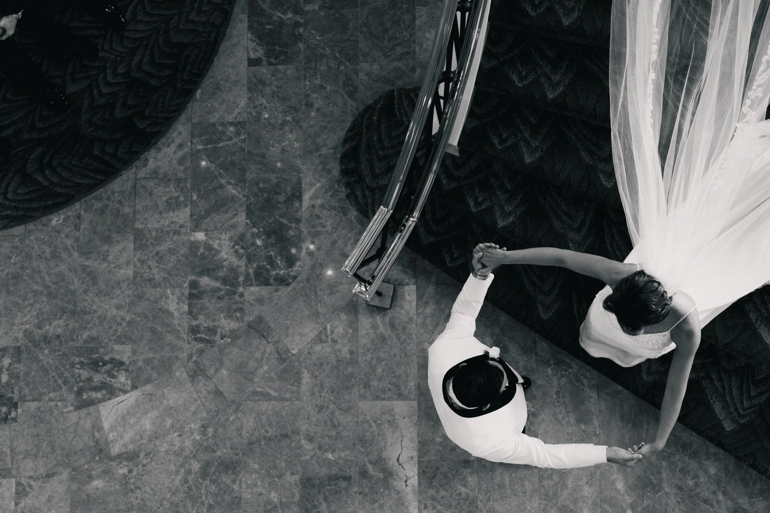 A woman in a white dress with lace details and a woman in white pants and a white shirt holding hands in a dance pose on a dark marble floor with a mirror and a black patterned carpet.
