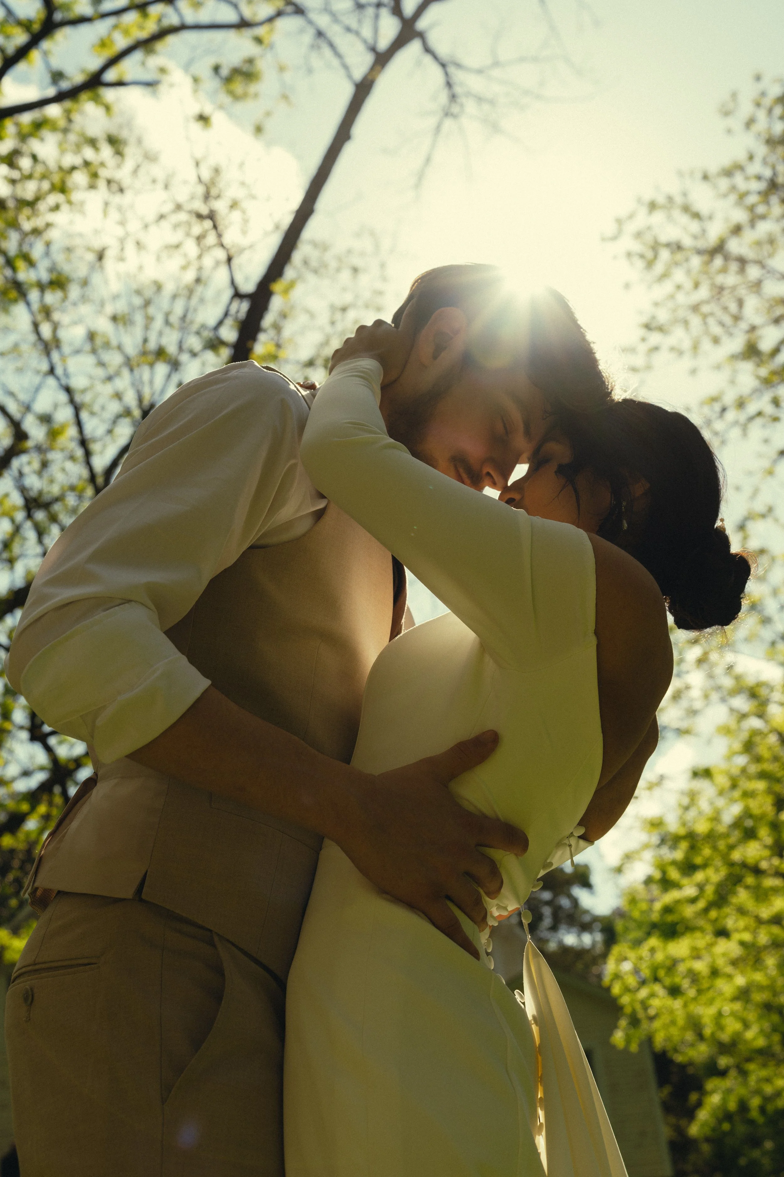 A couple sharing a kiss outdoors during daylight, with the sun shining behind them and trees in the background.