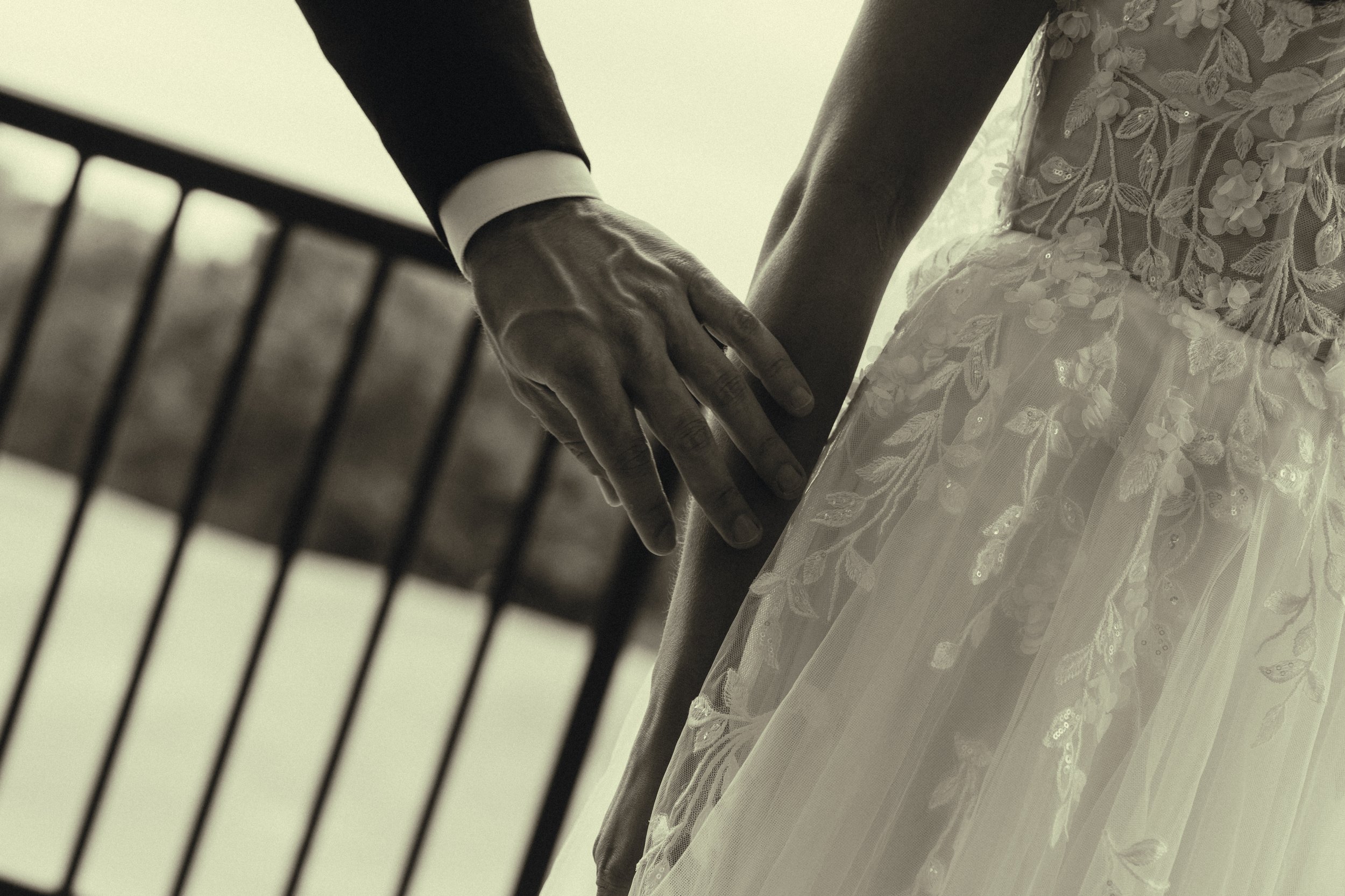Close-up of a bride and groom holding hands, with the bride wearing a lace wedding dress and the groom in a suit, as seen through a decorative fence.