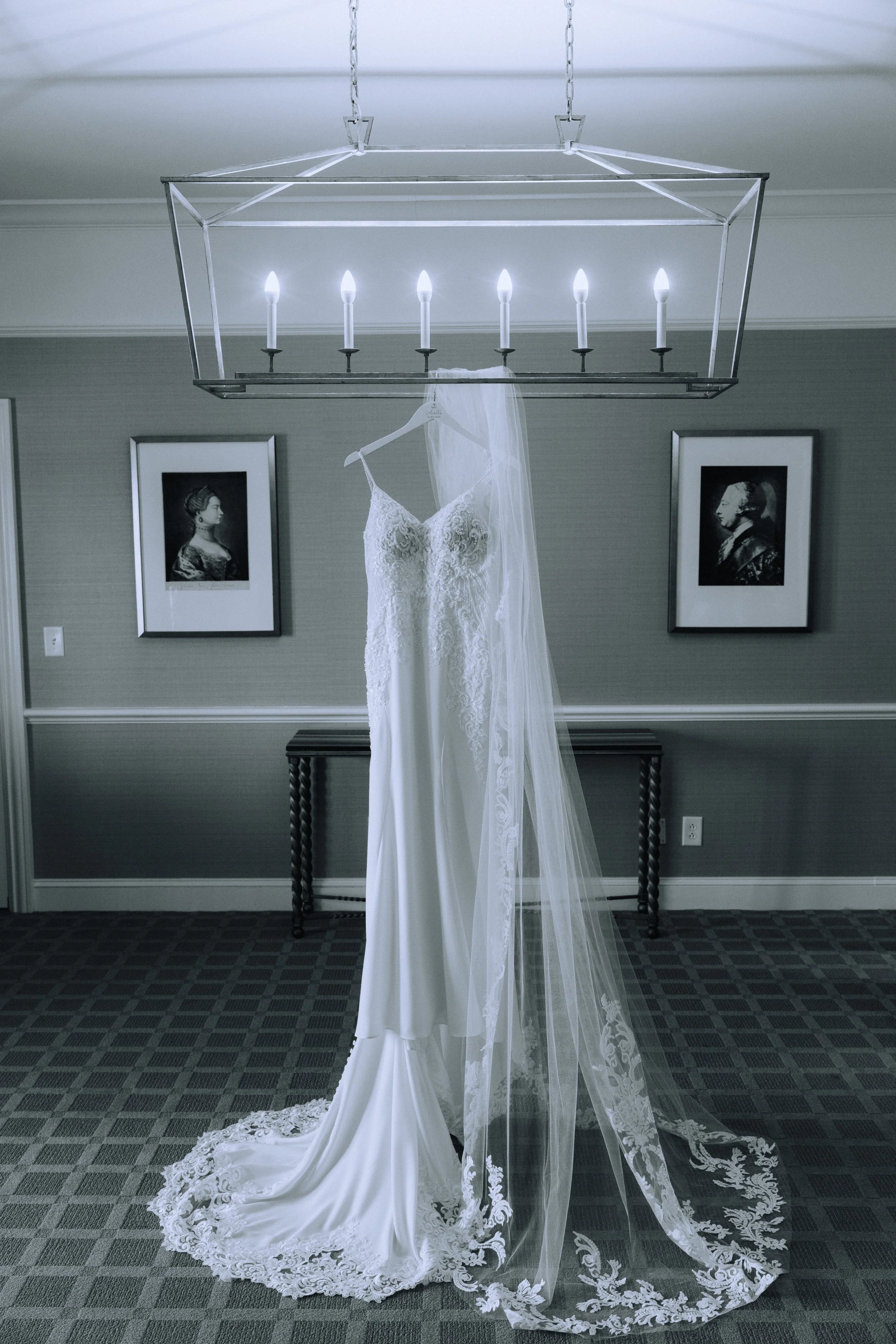 A wedding dress hanging on a hanger beneath a modern rectangular chandelier with candles, with framed black-and-white portraits on a gray wall behind it.
