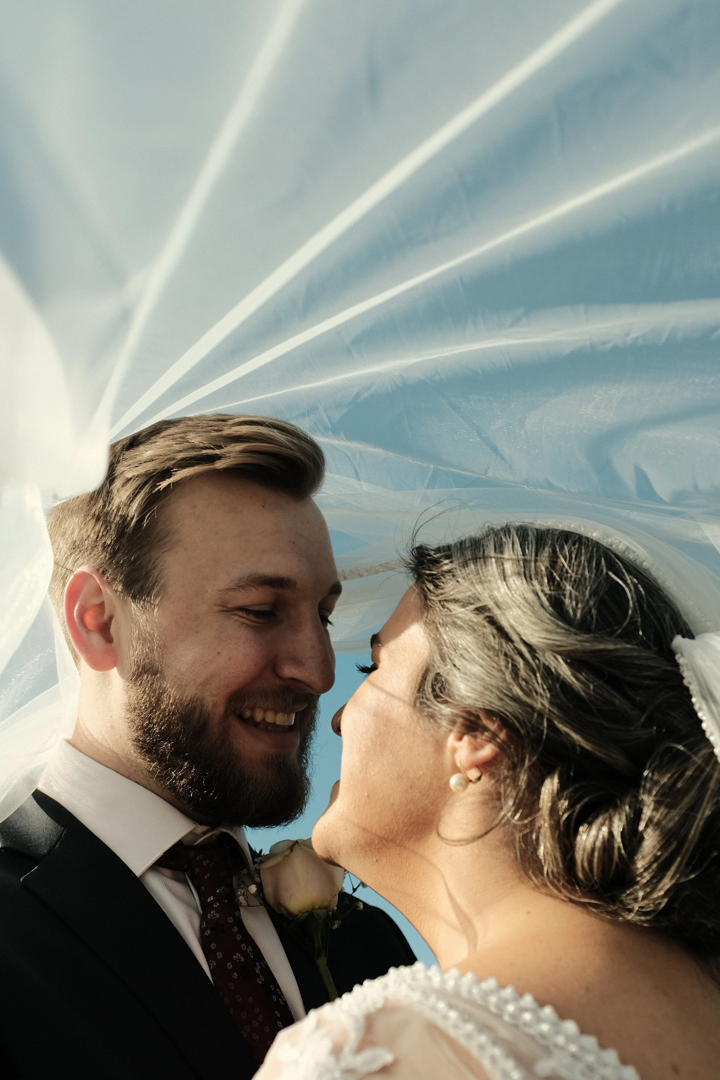 A close-up of a happy couple, a man with a beard in formal attire and a woman with earrings, sharing a kiss under a white veil outdoors.