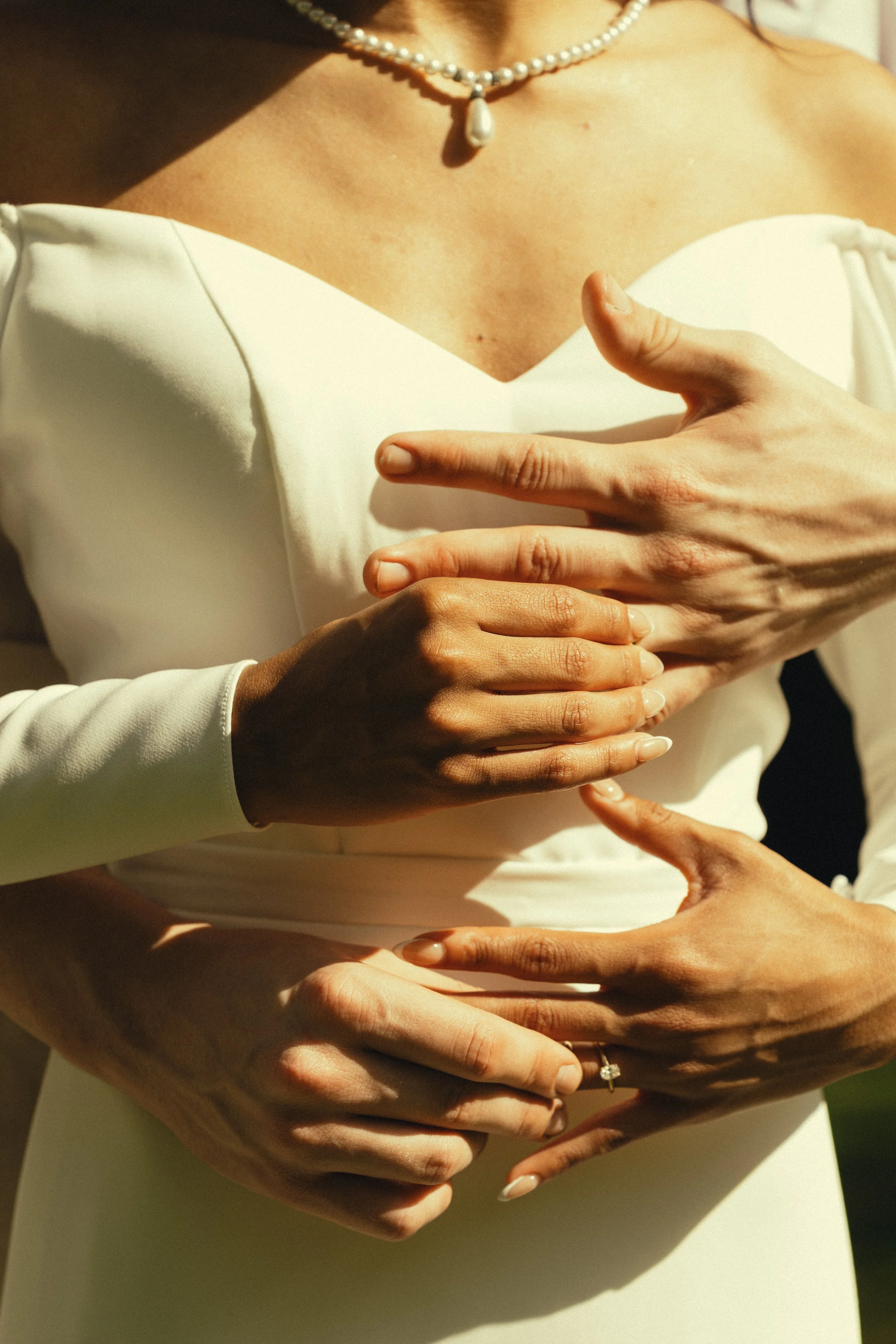 A woman wearing a white dress with a pearl necklace, with multiple hands touching her stomach area.