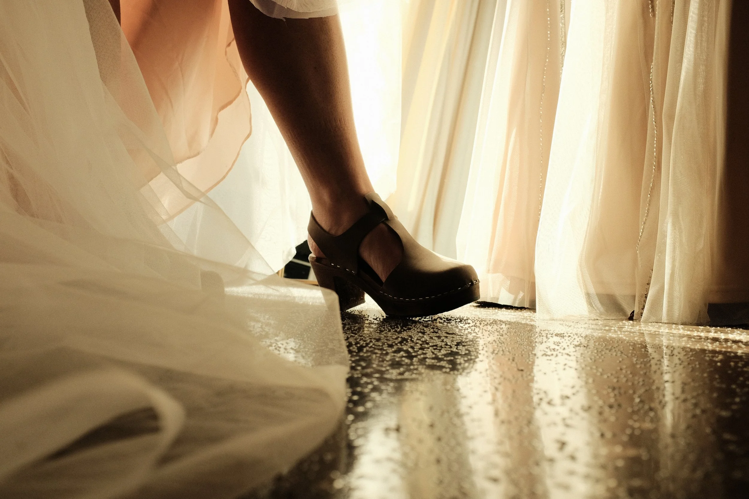 Close-up of a person's leg wearing black high-heeled shoes, standing on a carpet near a sheer curtain, with sunlight filtering through.