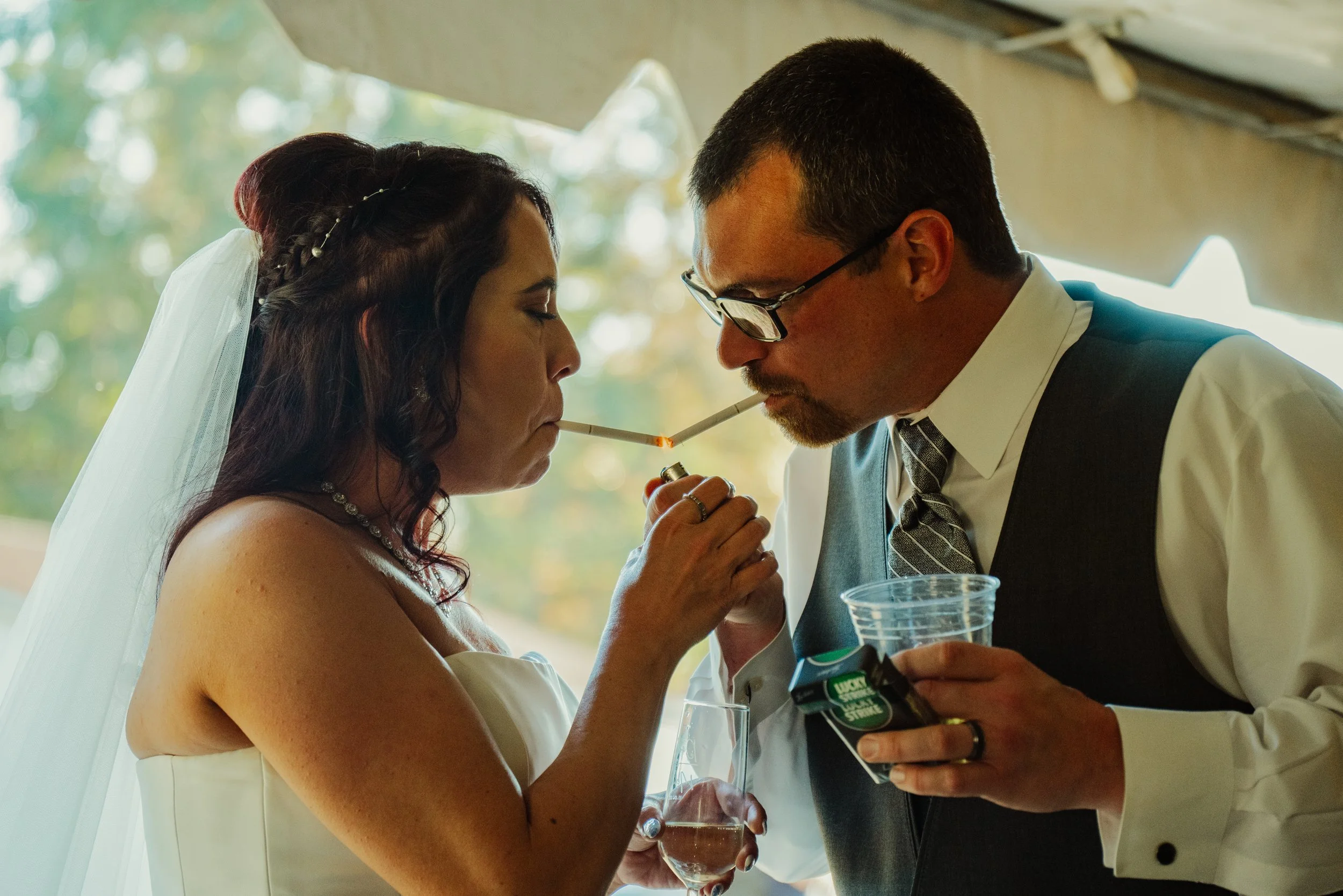 A bride and groom with the bride wearing a white wedding dress and veil and the groom in a suit with a tie, sharing a moment with a lit cigarette during their wedding reception.