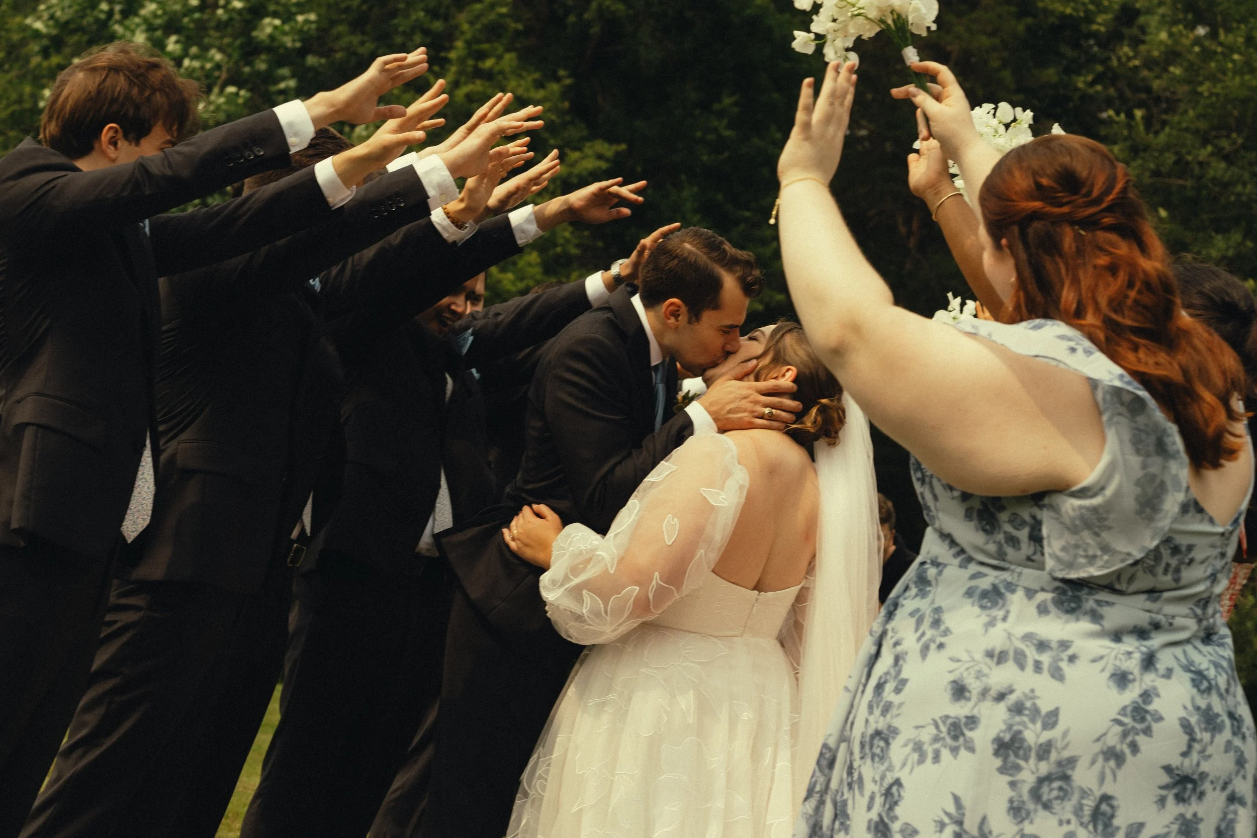 A wedding ceremony where a bride and groom are kissing while surrounded by friends and family, with arms reaching out to the couple and holding flowers.