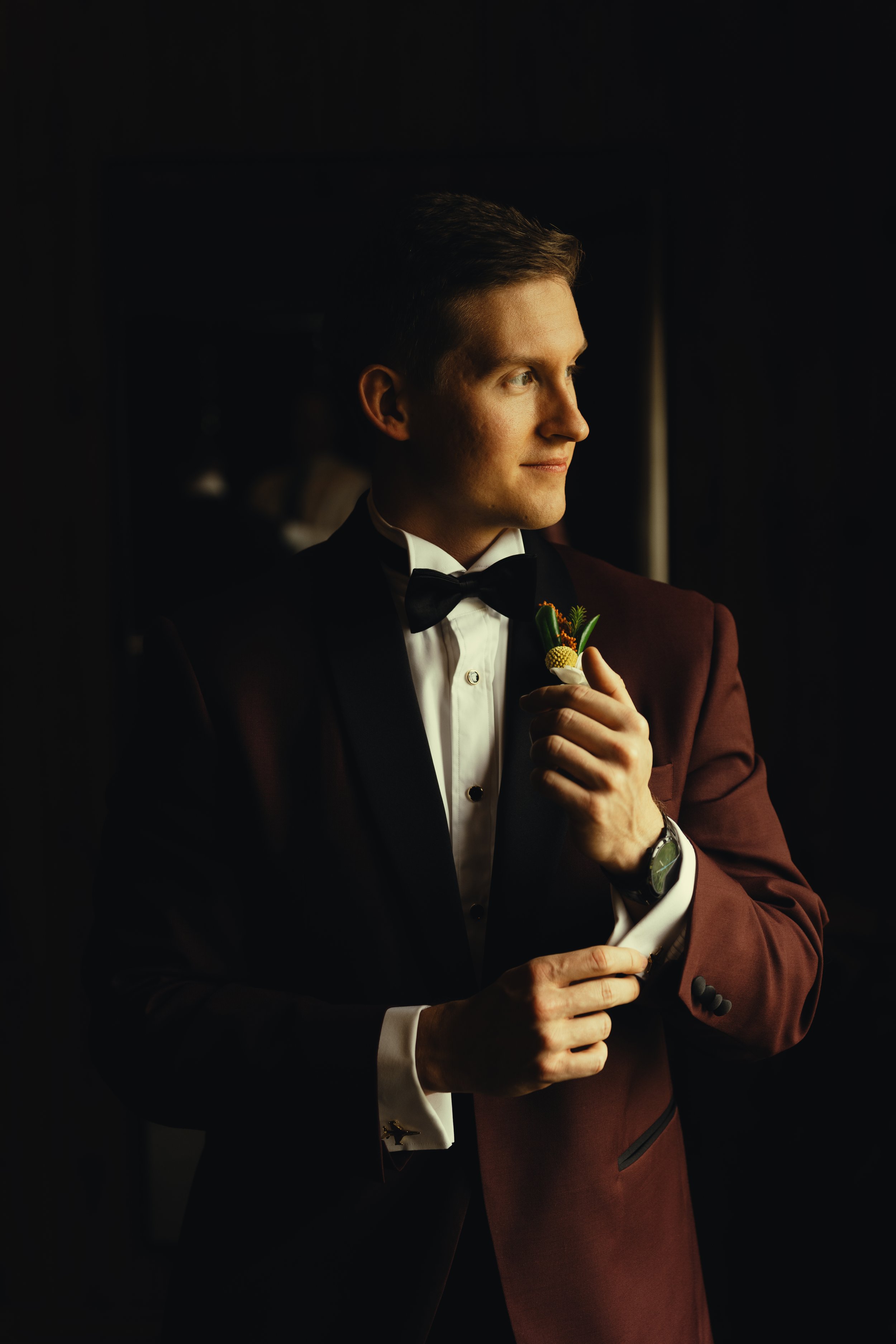 A young man in formal wear, including a tuxedo with a bow tie, holding a telephone receiver to his ear and smiling slightly. He is standing against a dark background with soft lighting on his face.