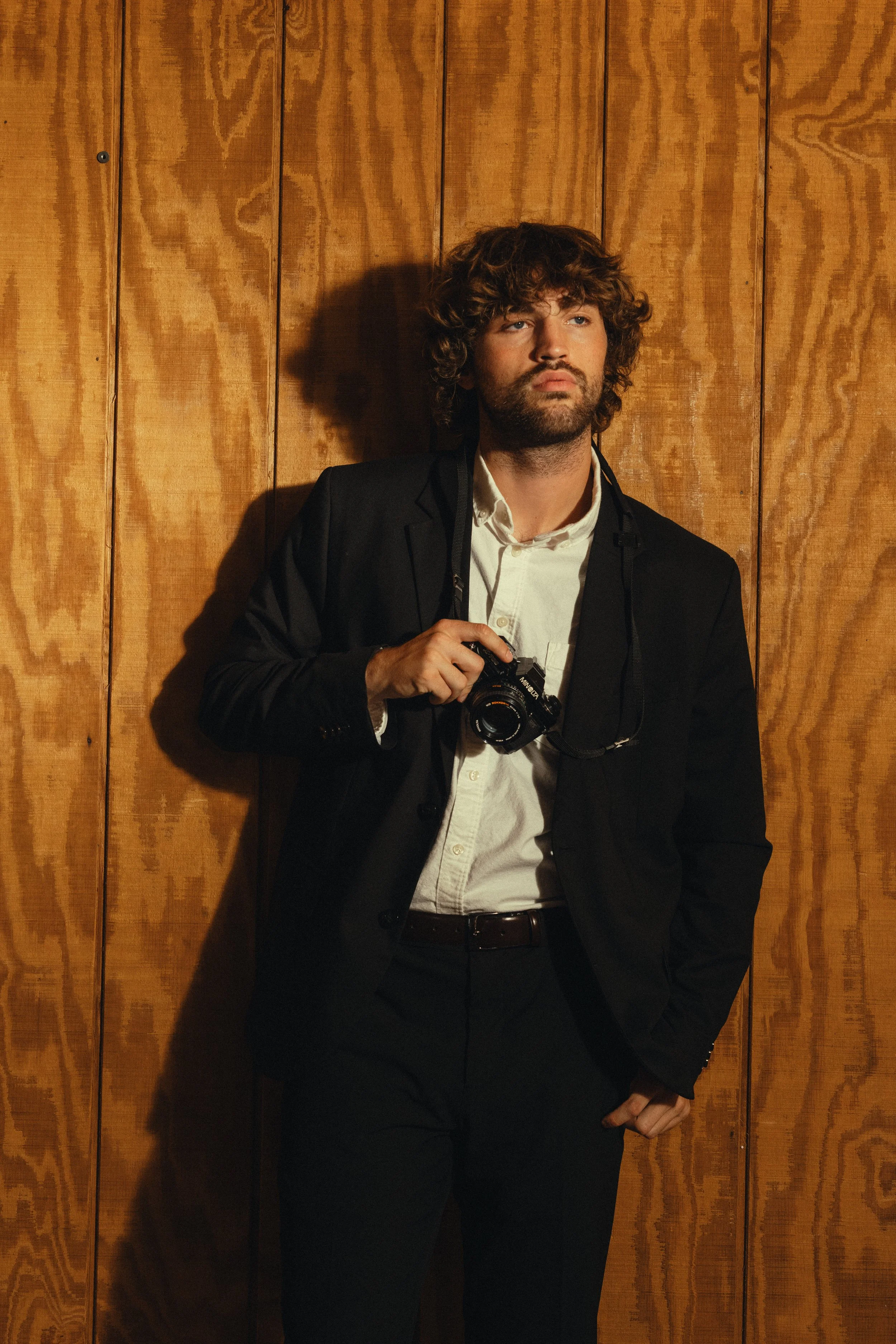 A man with curly brown hair and a beard wearing a black suit jacket, white shirt, and holding a camera, standing against a wooden wall.