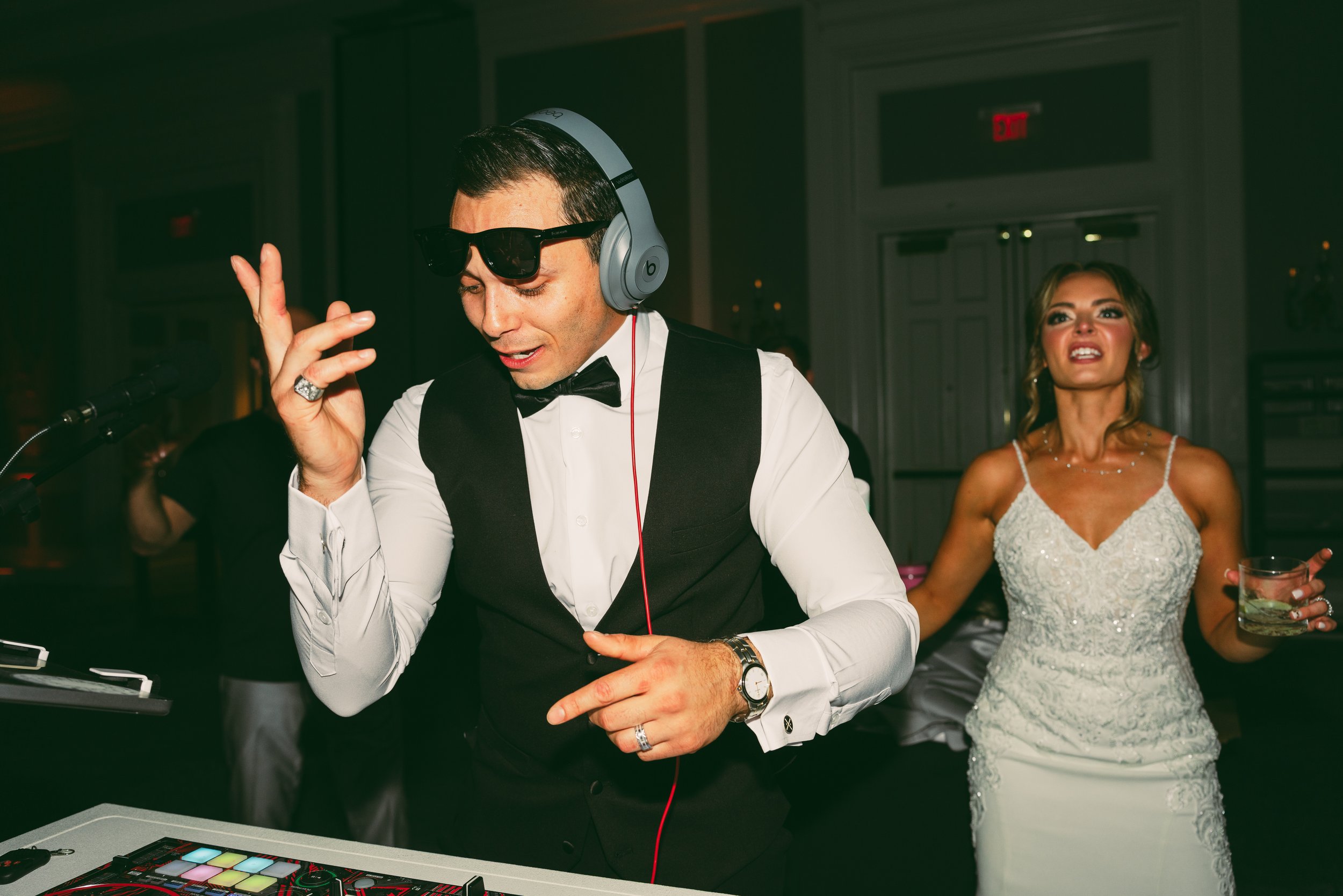 A man in a tuxedo shirt, black vest, and bow tie, wearing sunglasses and headphones, appears to be DJing at a party. Behind him, a woman in a white lace dress with makeup and styled hair is holding a drink, looking and speaking with an expressive fac