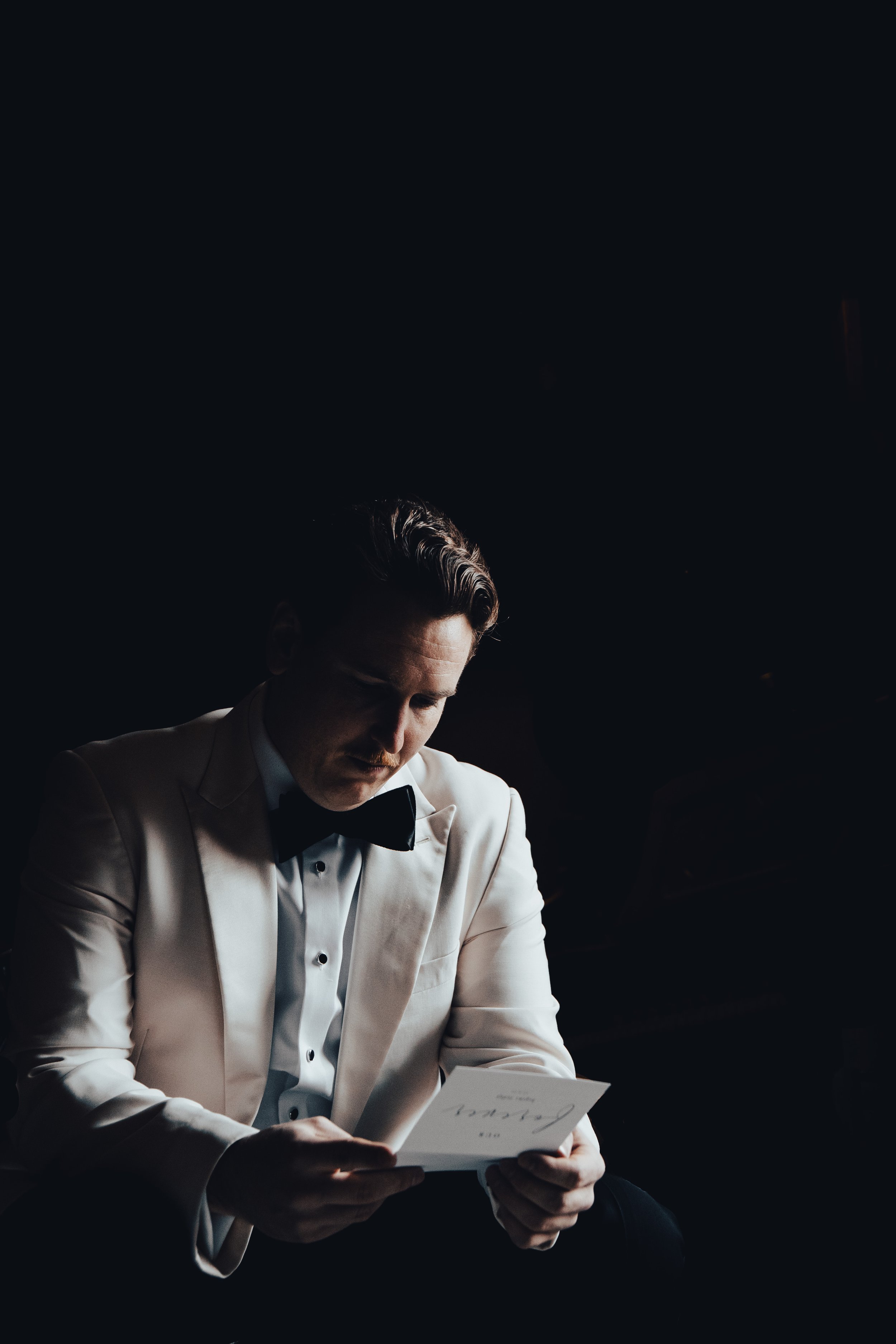 A man in a white tuxedo with a black bow tie is sitting in a dark room, reading a letter or card.