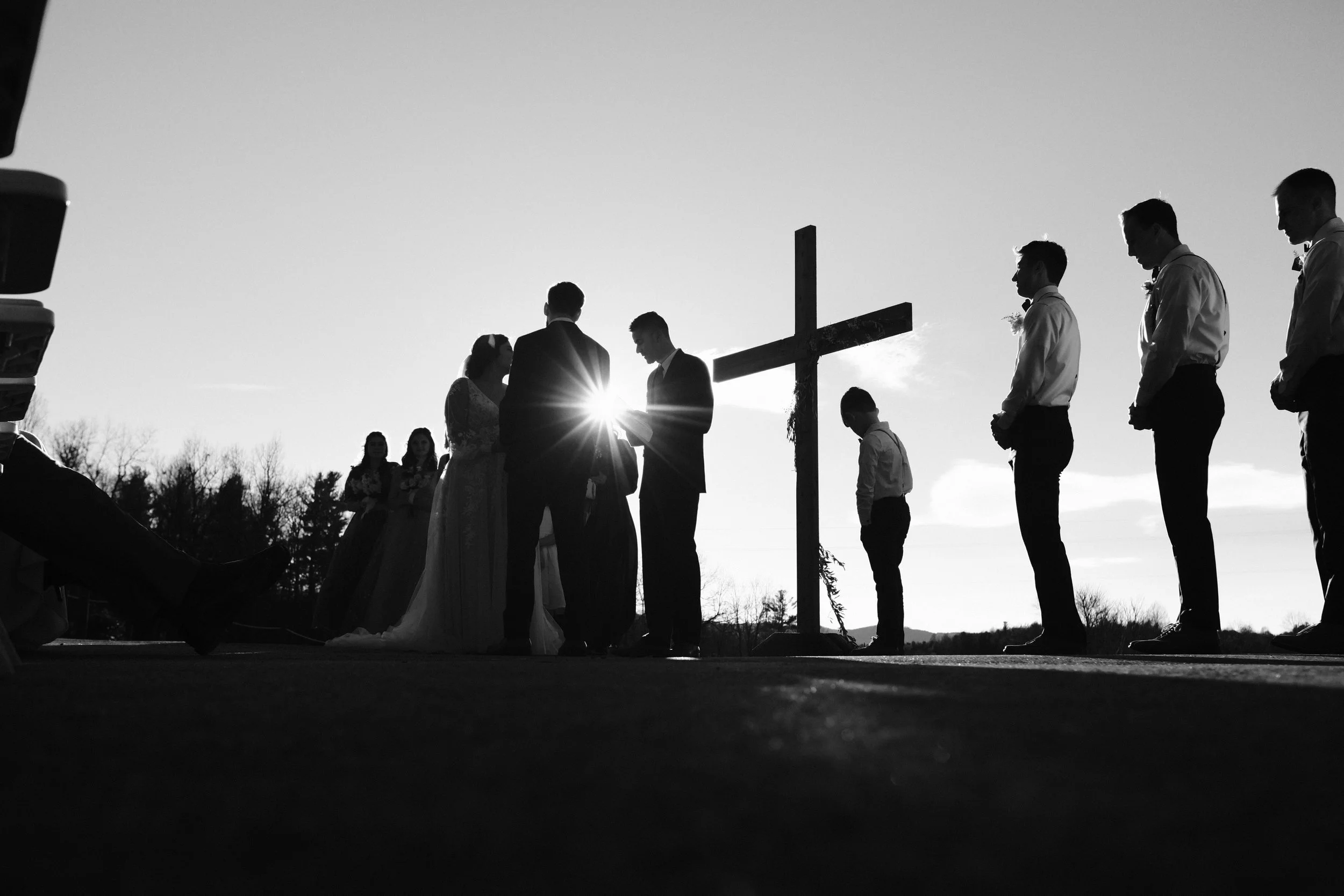 Silhouette of a wedding ceremony outdoors at sunset with a cross, bride and groom exchanging vows, and guests standing nearby.