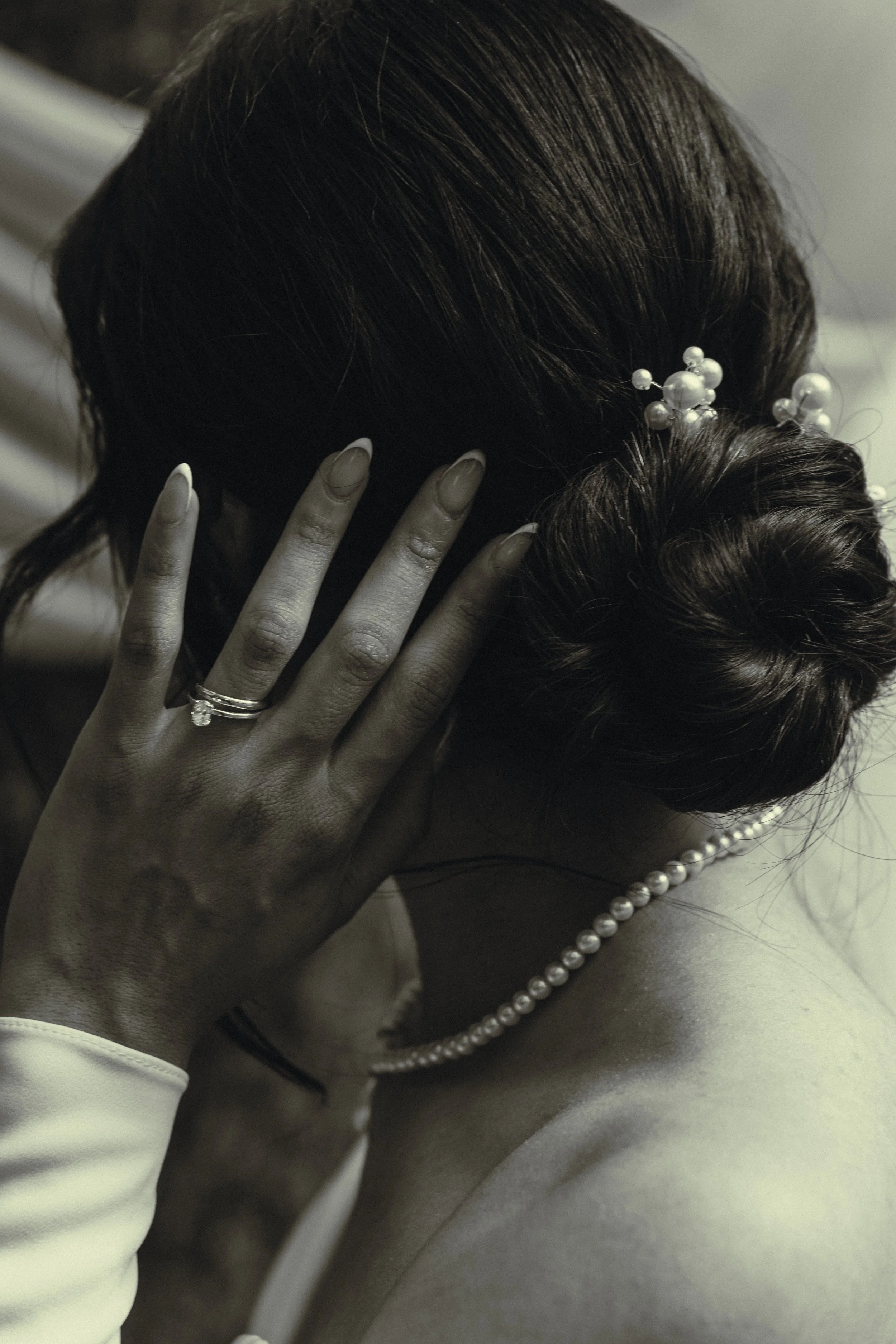 Close-up of a woman wearing a pearl necklace and pearl hair accessories, with her hand resting on her face, her hair styled in an elegant updo, in a black and white toned photograph.