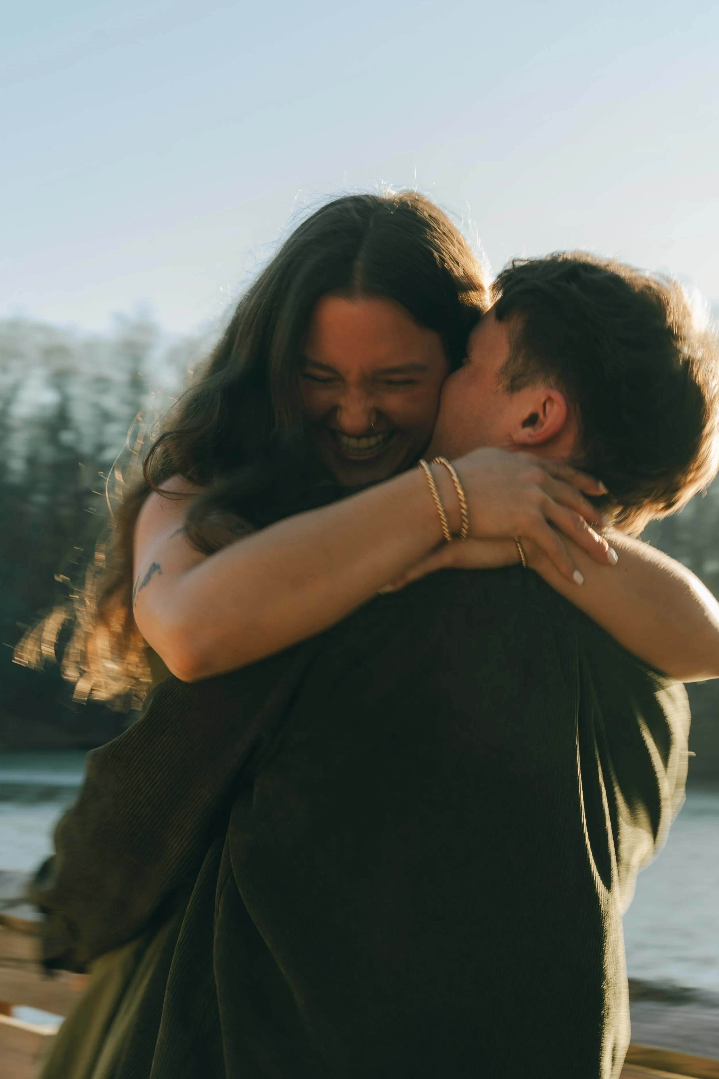 A person with long hair and jewelry hugging another person outdoors near water with buildings in the background.