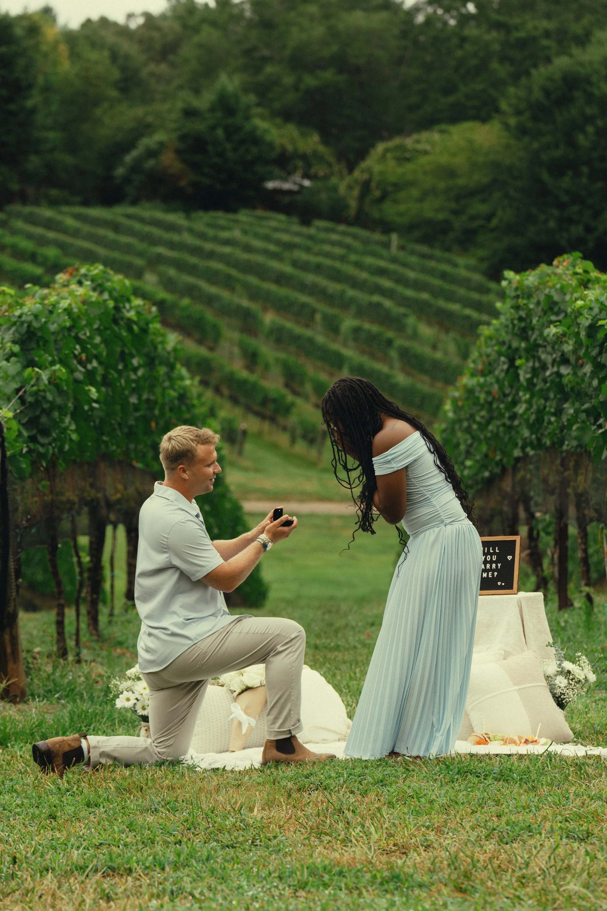 A man proposing marriage to a woman in a vineyard, with the man kneeling on one knee and the woman bowing her head, surrounded by green grapevines and outdoor wedding decor.