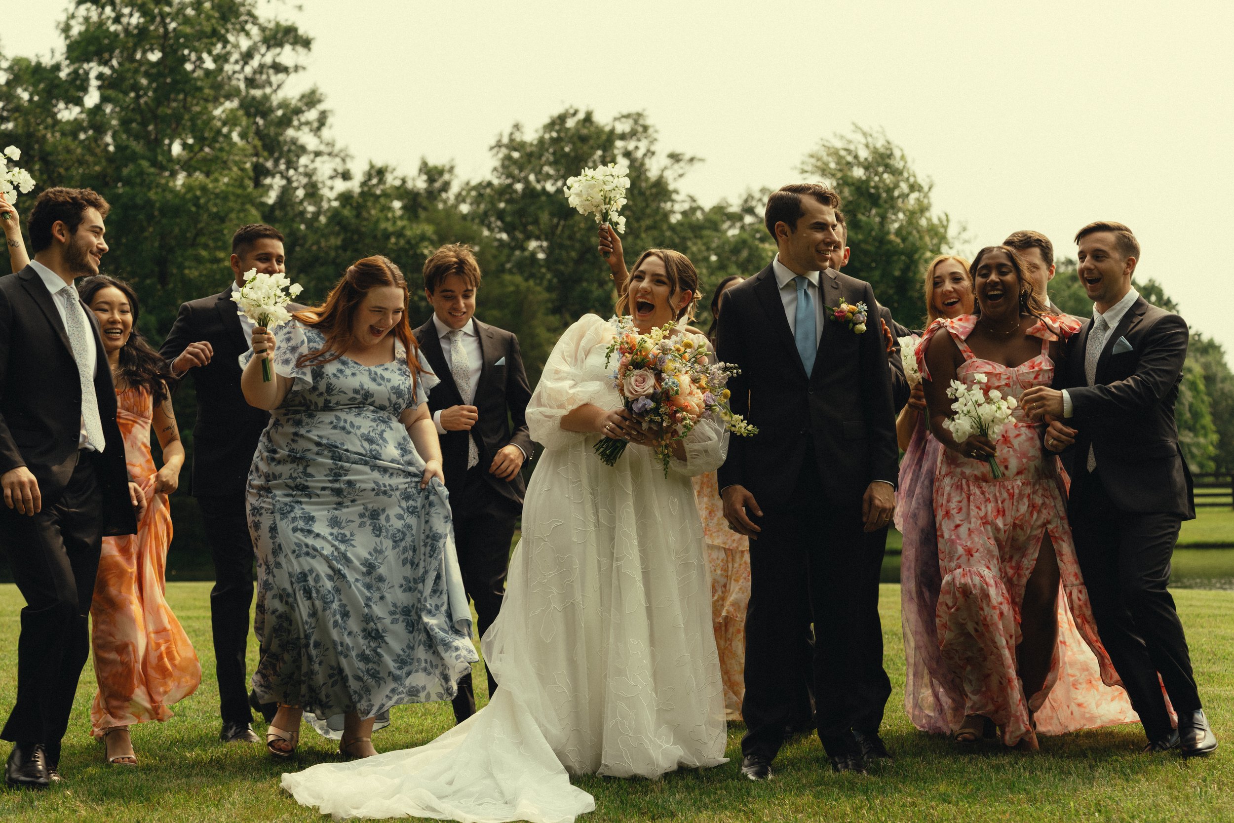 A group of people celebrating a wedding outdoors, with the bride in a white dress holding a bouquet and others holding flowers, all smiling and walking on grass.