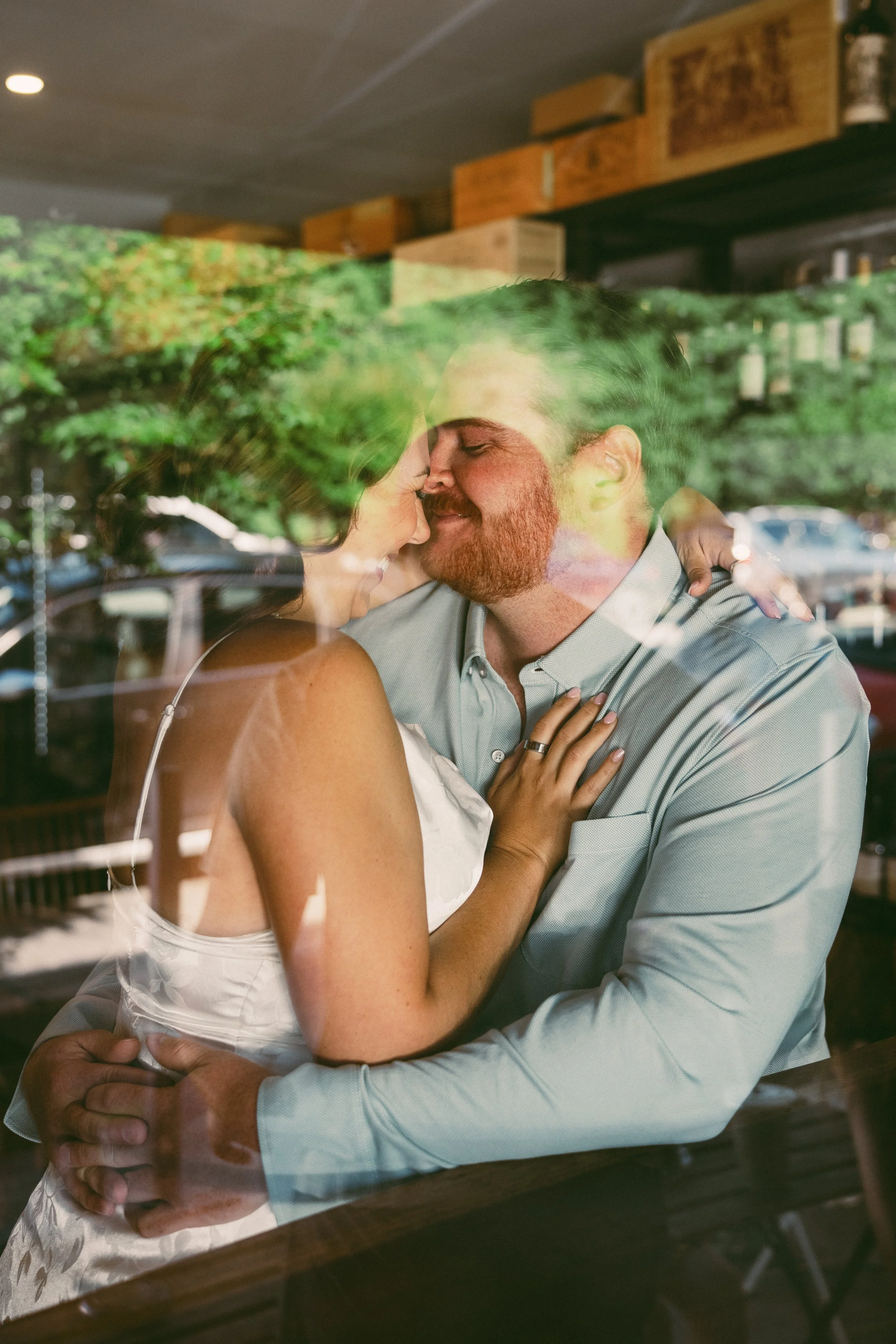A man and woman embrace and smile inside a restaurant, with their reflection visible in a window.