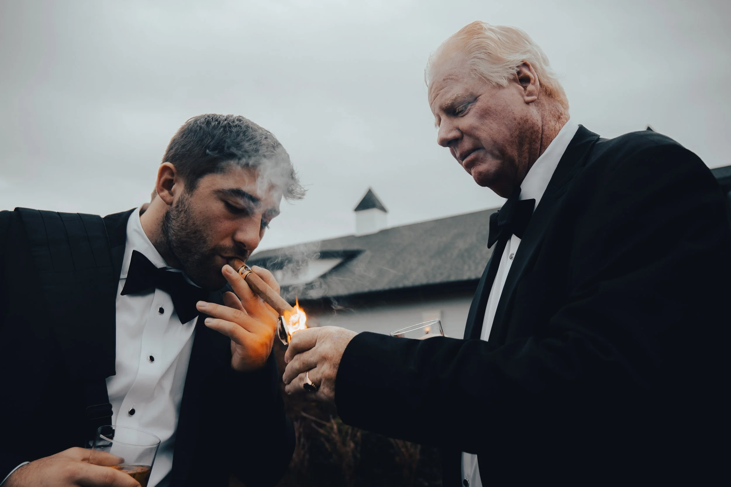 Two men in tuxedos, one older with white hair and the other younger with dark hair, are outside during daytime. The younger man is smoking a cigar while the older man is holding a glass of drink, and they are closely looking at each other. The backdr