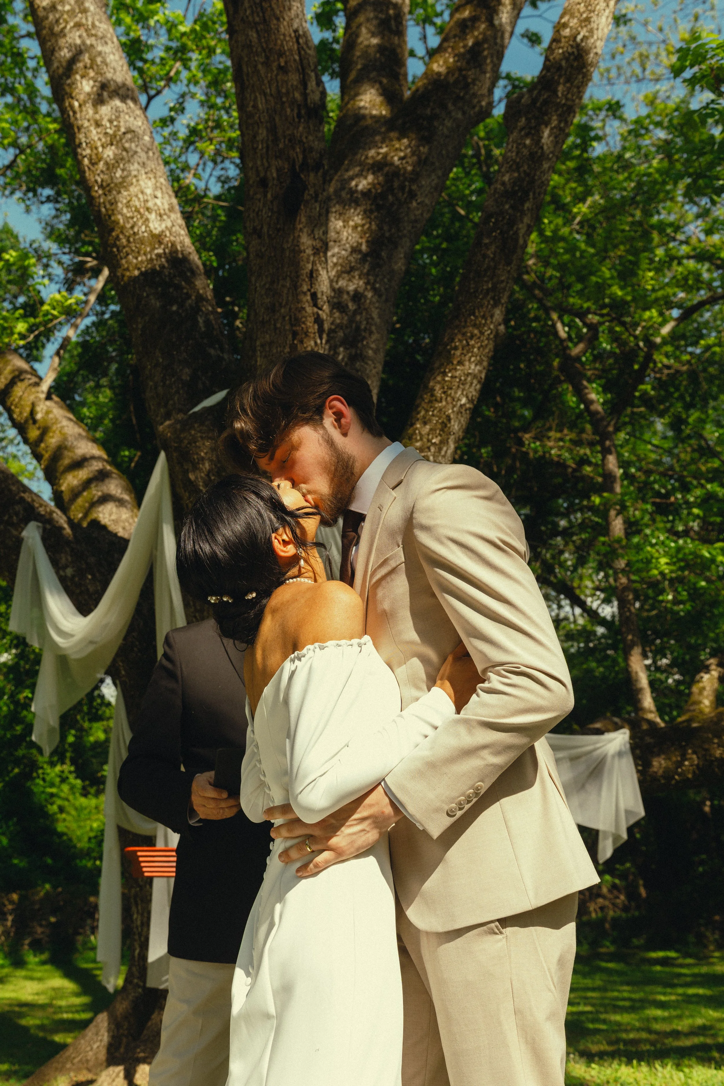 A couple sharing a kiss during their outdoor wedding ceremony, with a large tree and green foliage in the background.