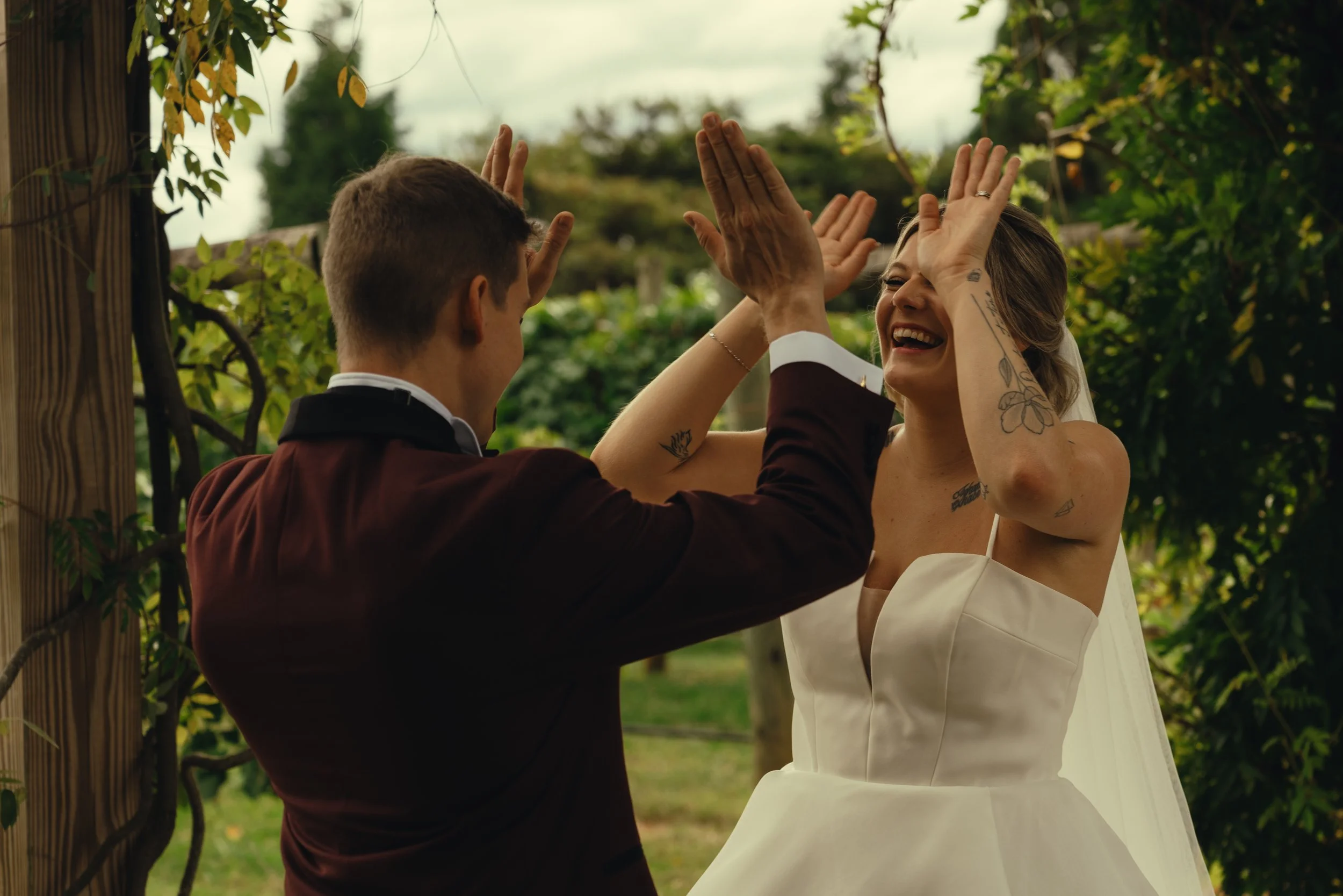 A couple celebrates with a high five in an outdoor garden setting during their wedding. The woman is wearing a white wedding dress and the man a dark suit. Both are smiling joyfully, and the woman has tattoos on her arms.
