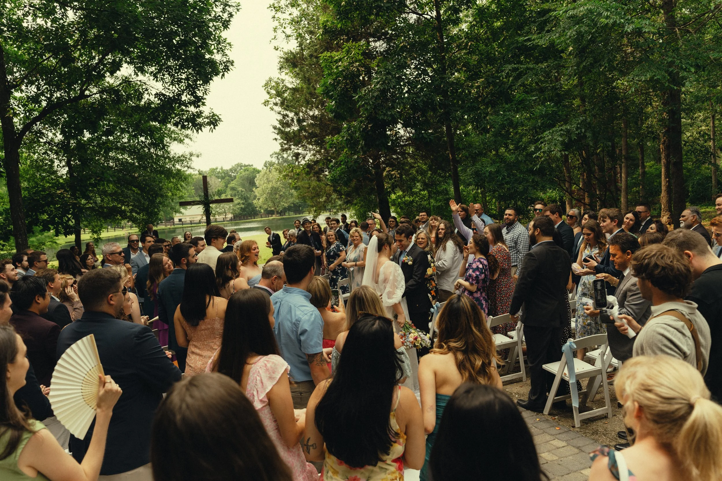 Crowd of people gathered outdoors for a wedding ceremony in a park with trees and a cross in the background.