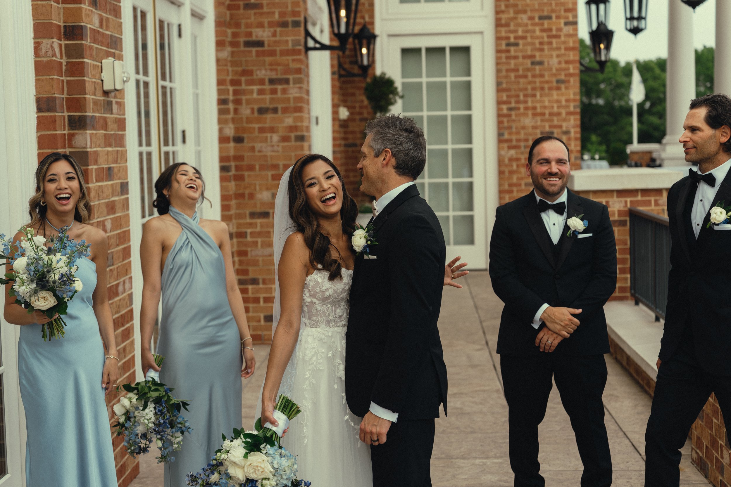 A wedding scene outside a brick building with seven people, including the bride and groom, dressed in formal attire and smiling. The bride is in a white wedding dress holding a bouquet, and the groom is in a black tuxedo. Bridesmaids and groomsmen ar