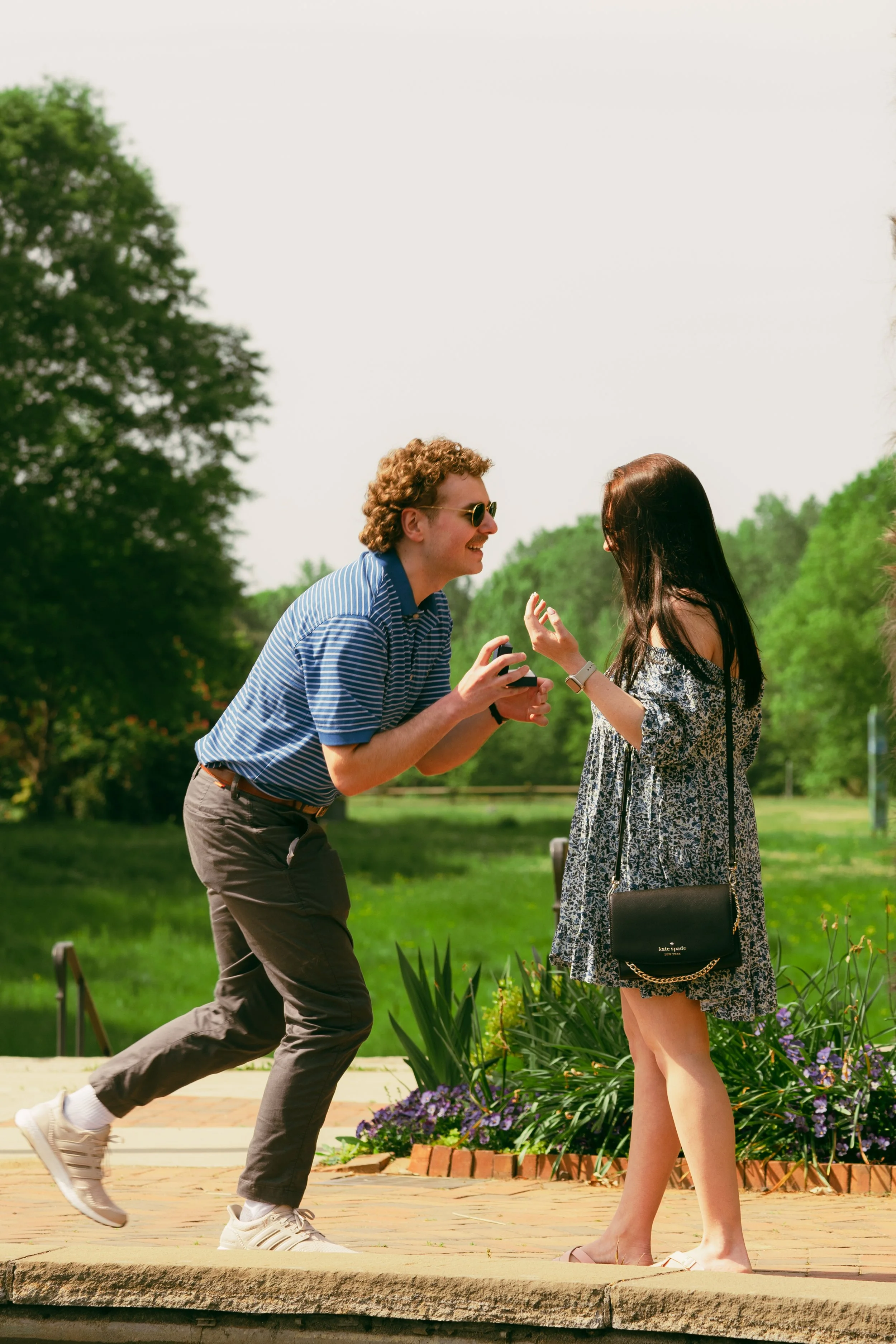 A man in a blue striped shirt and gray pants is proposing marriage to a woman in a floral dress outdoors in a park with trees and flowers, while a man in a blue striped shirt is kneeling.