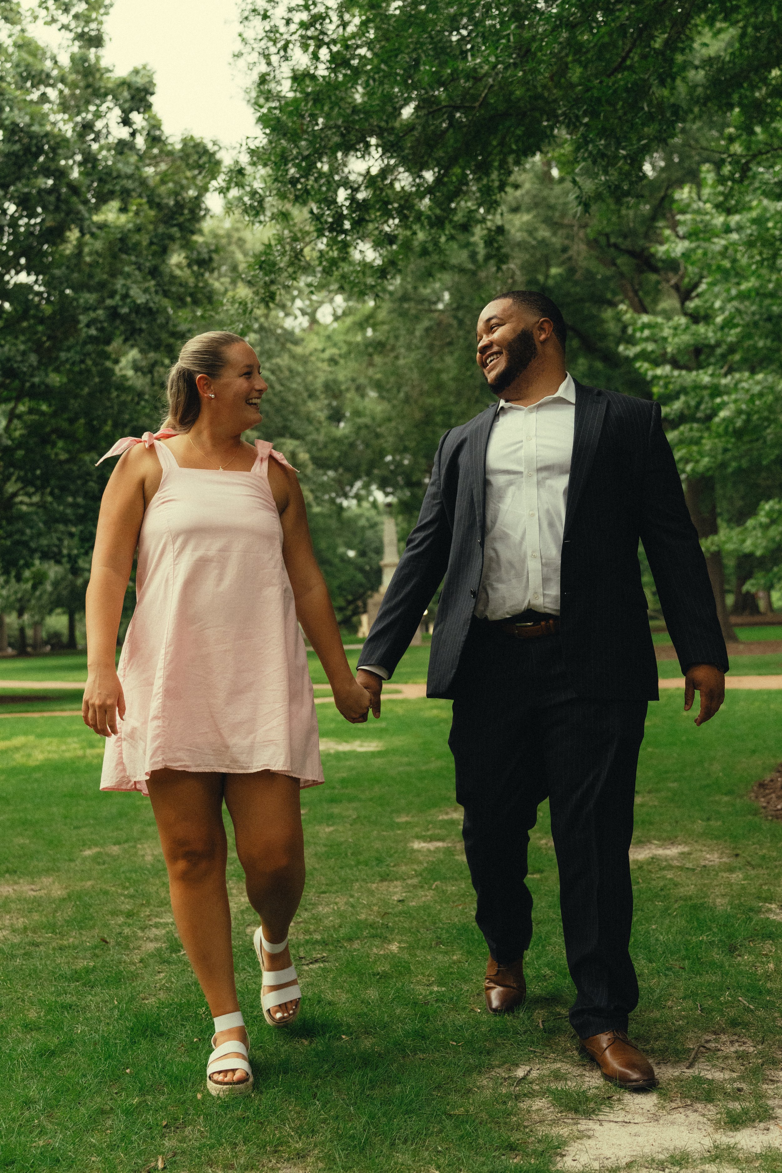 A smiling couple holding hands and walking in a park, surrounded by green trees.