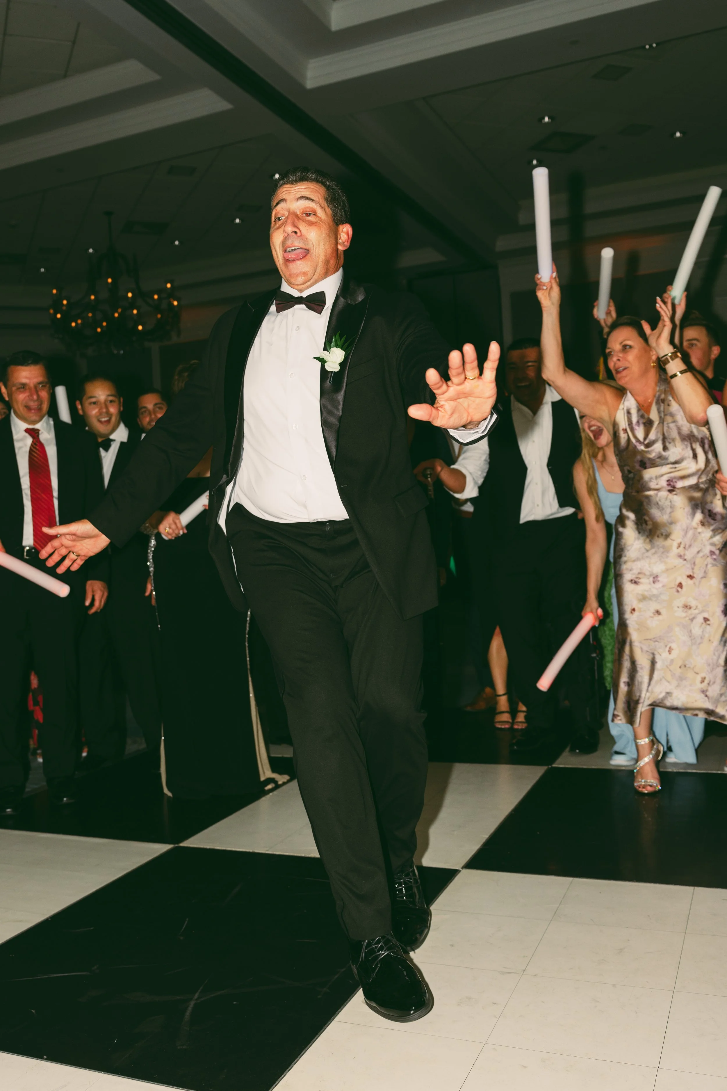 People dancing at a formal event, wearing elegant attire and holding glow sticks, with a man in a tuxedo in the foreground.